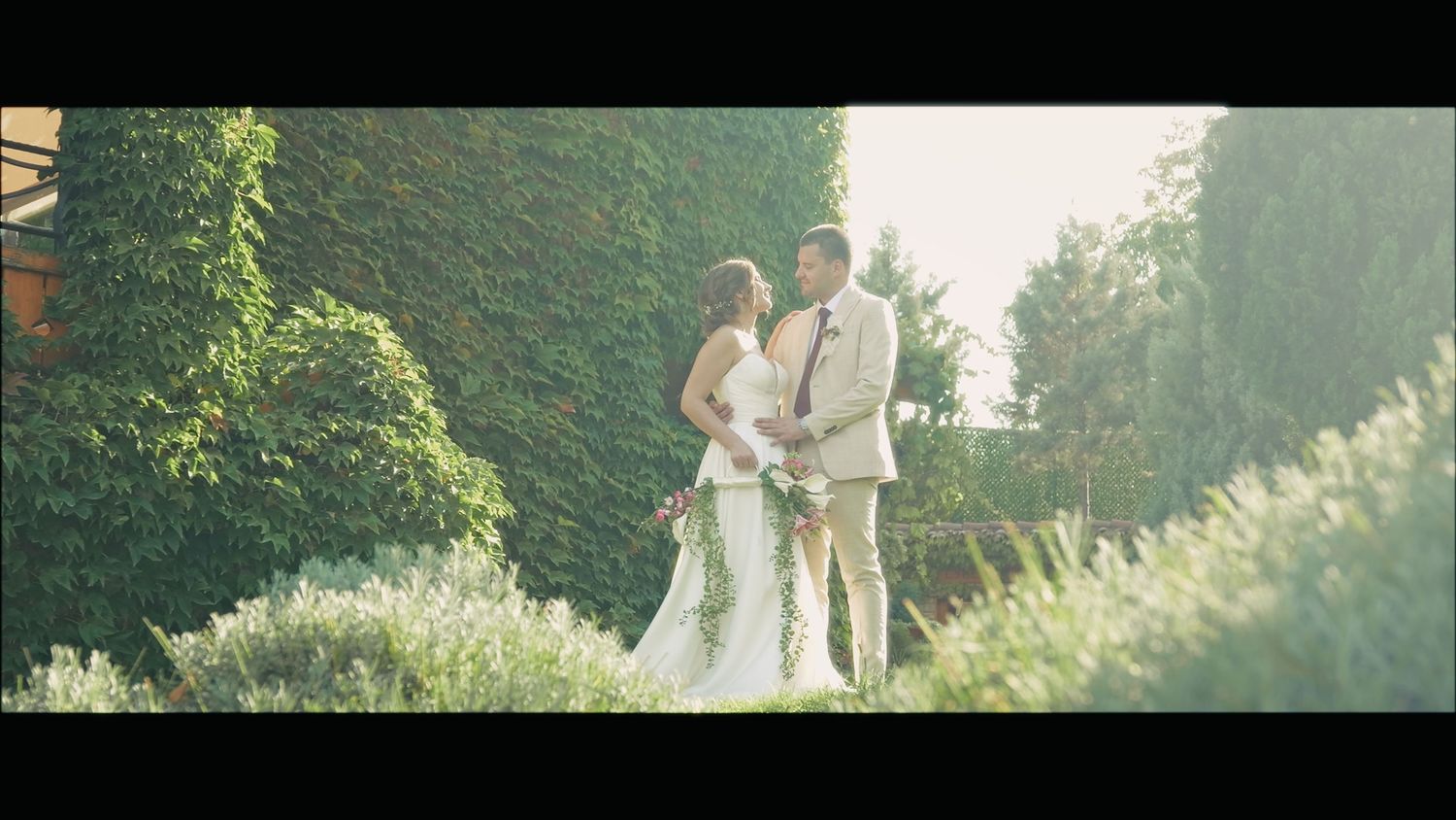 A couple poses in formal attire against a backdrop of green hedges and gardens.