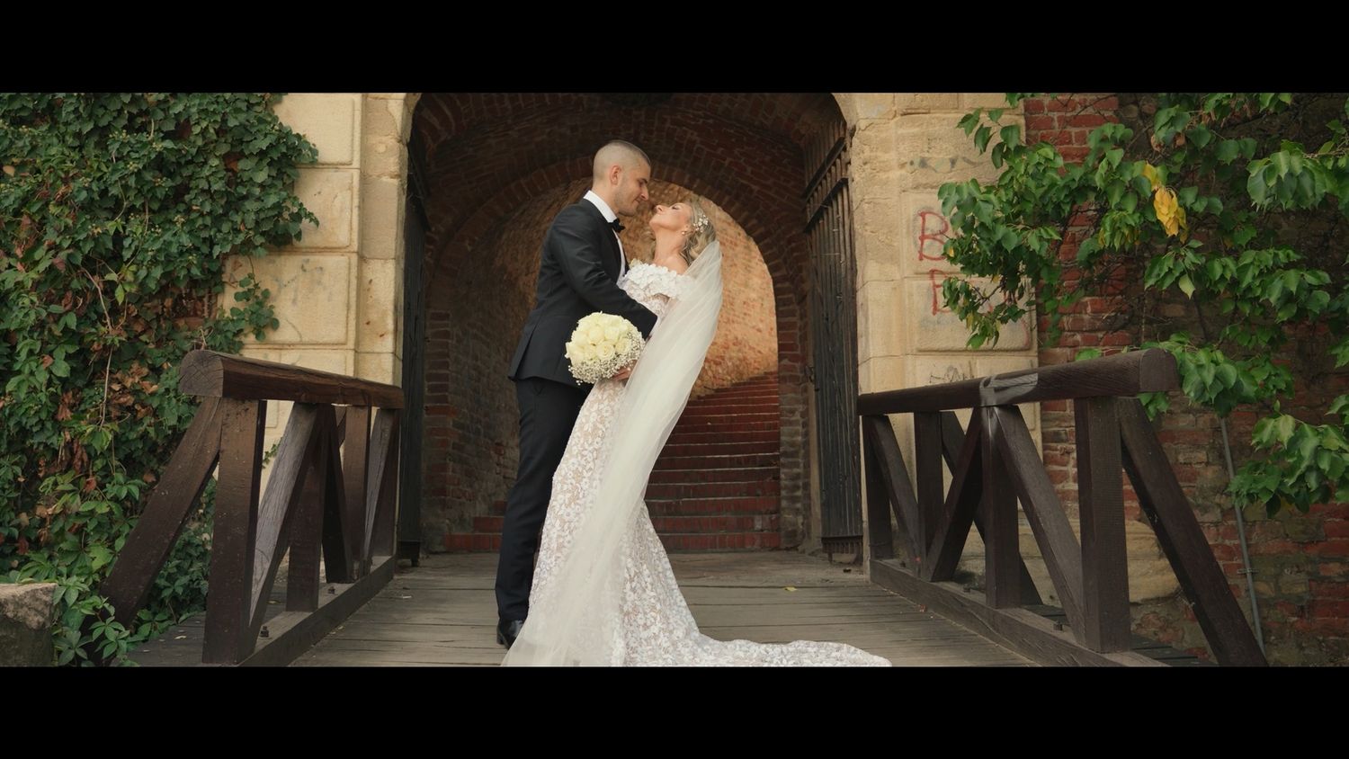 Wedding couple posing in front of rustic brick archway surrounded by greenery.