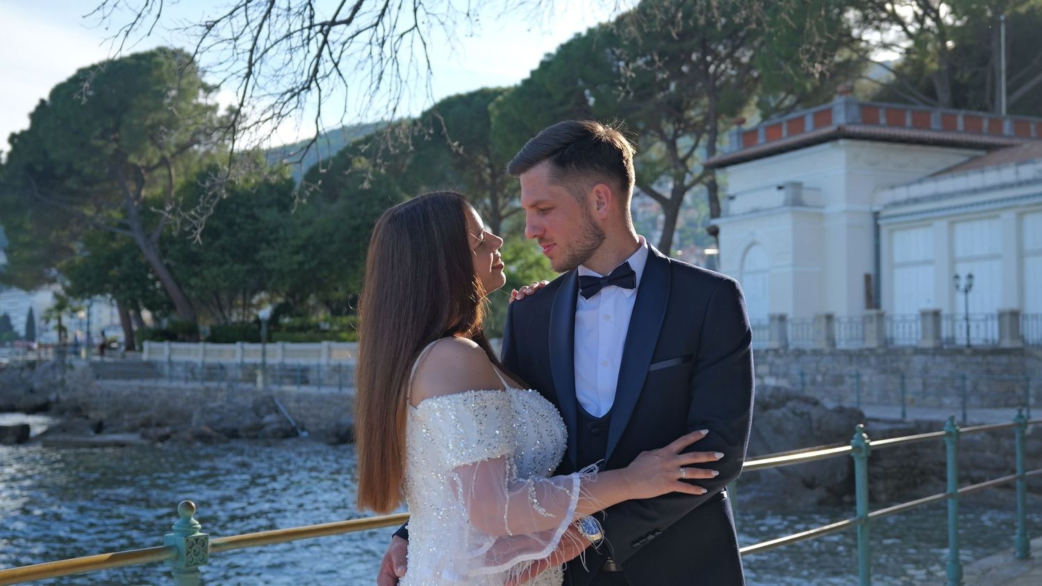 A couple in formal wedding attire shares a romantic moment by the waterfront at sunset.