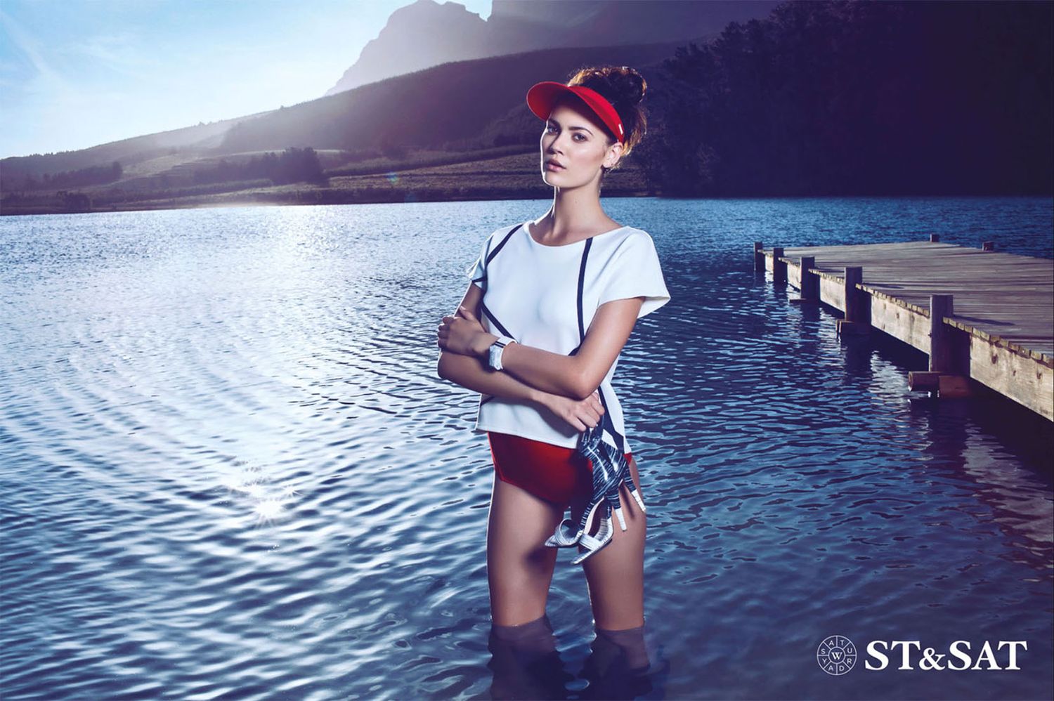 Fashion model poses on wooden dock by scenic mountain lake wearing casual summer outfit.