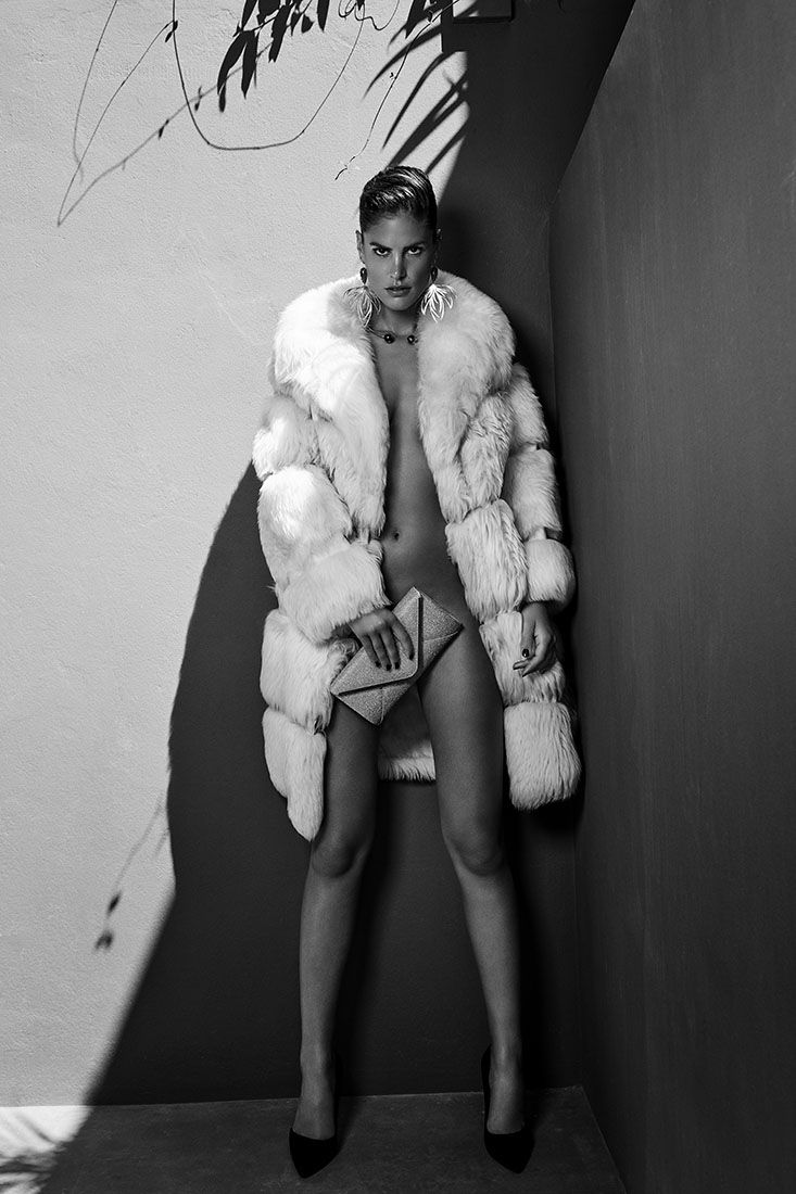 Fashion model wearing white fur vest poses against dark background in dramatic black and white photography series.