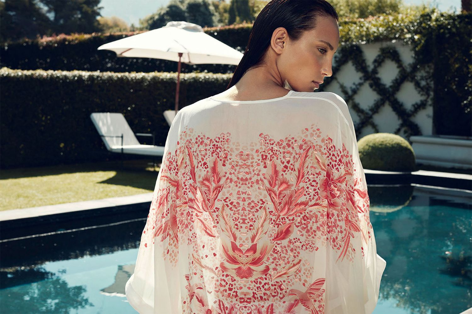 Woman in white kaftan with coral print relaxing by luxury pool.