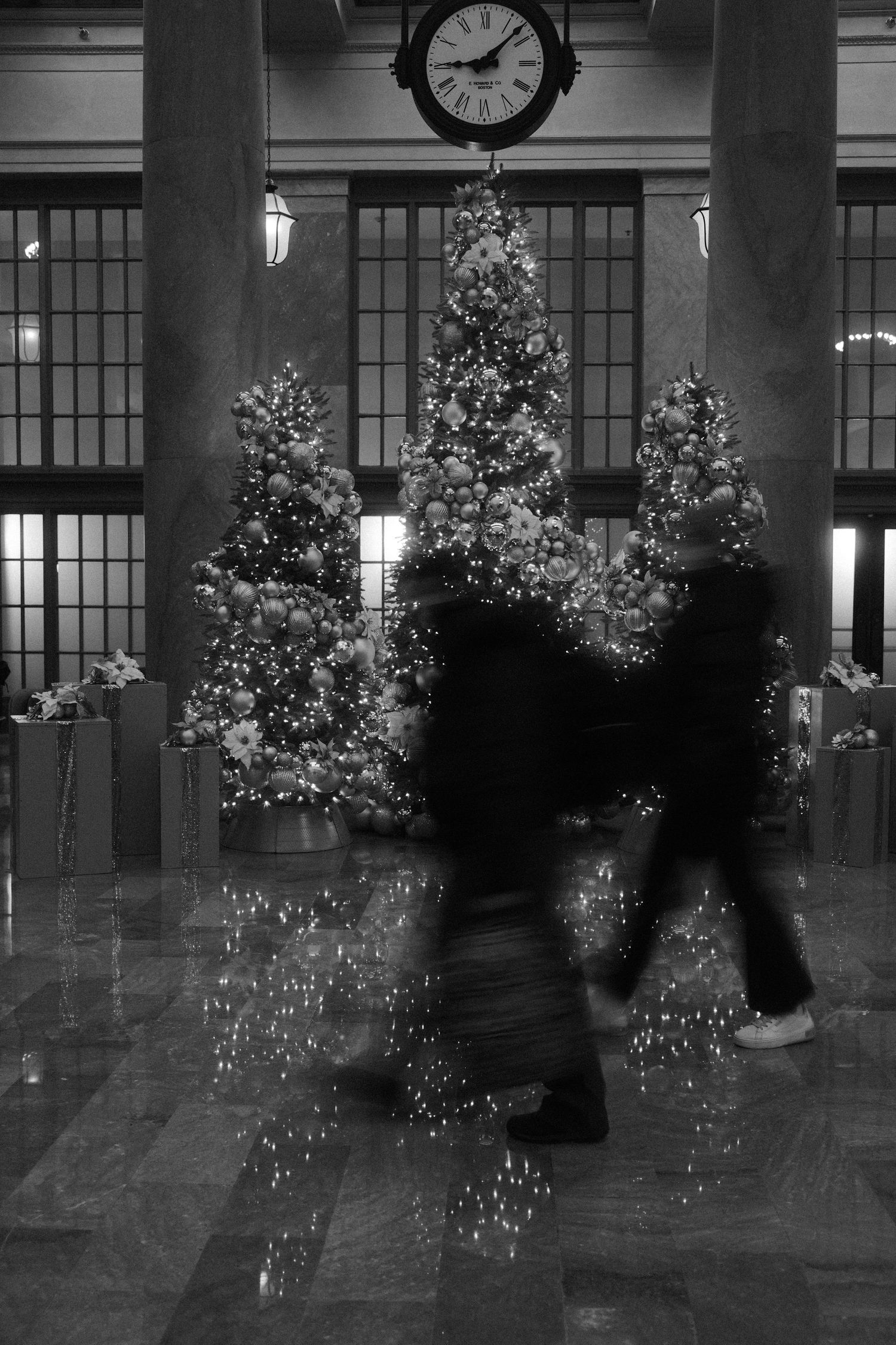 People walking past decorated Christmas trees inside a building with a large clock overhead.