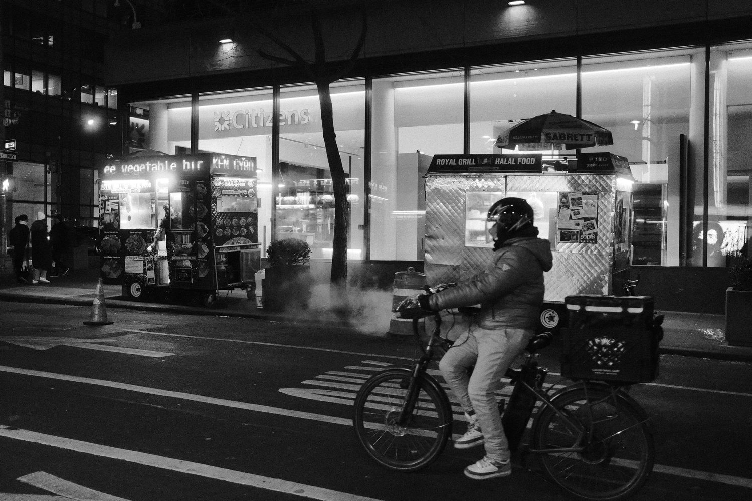 Cyclist with a delivery bag riding past food carts on a city street at night.