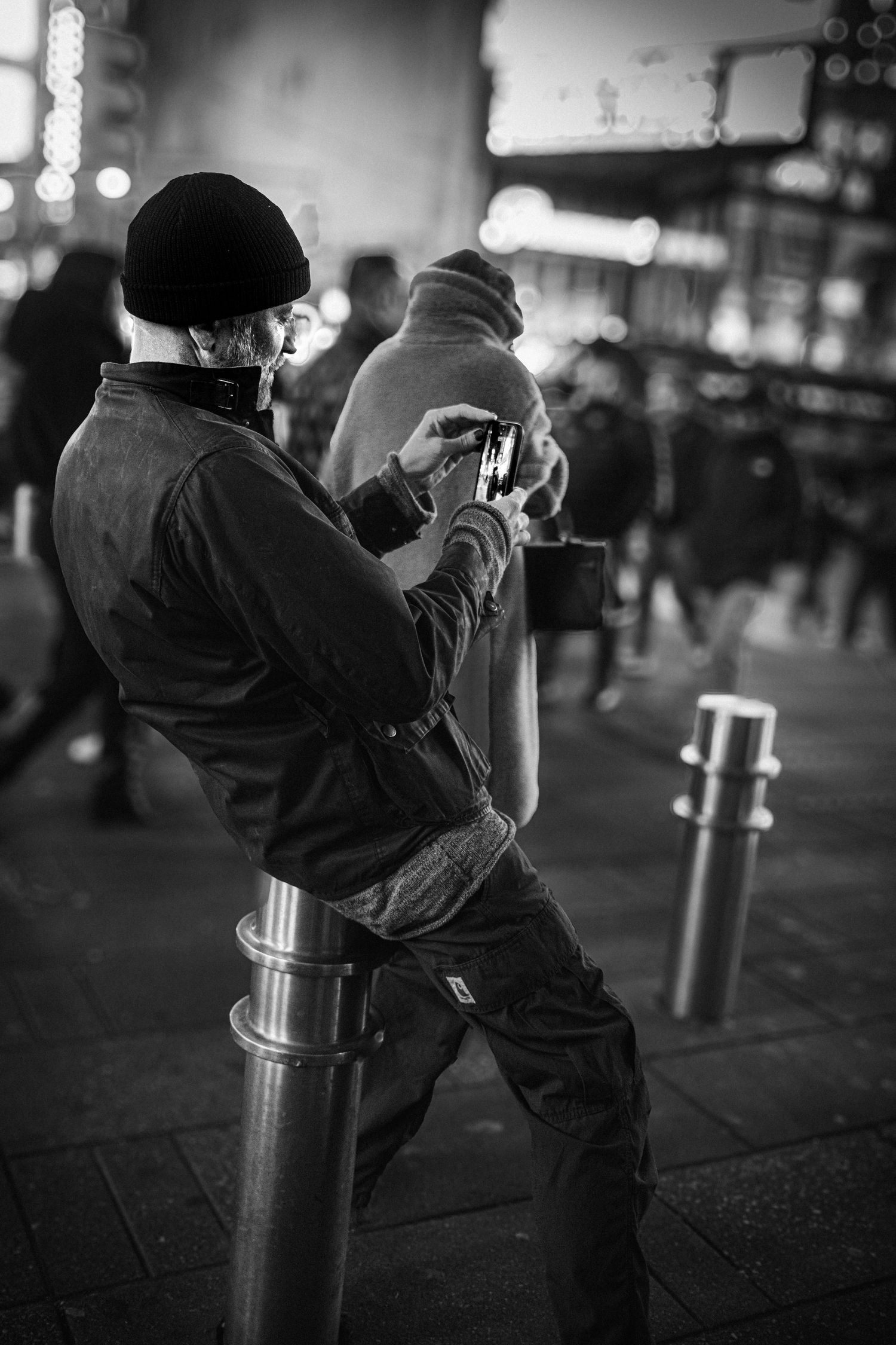 Man leaning on a metal bollard and looking at his phone in a crowded urban street at night. Man taking a photo on time square