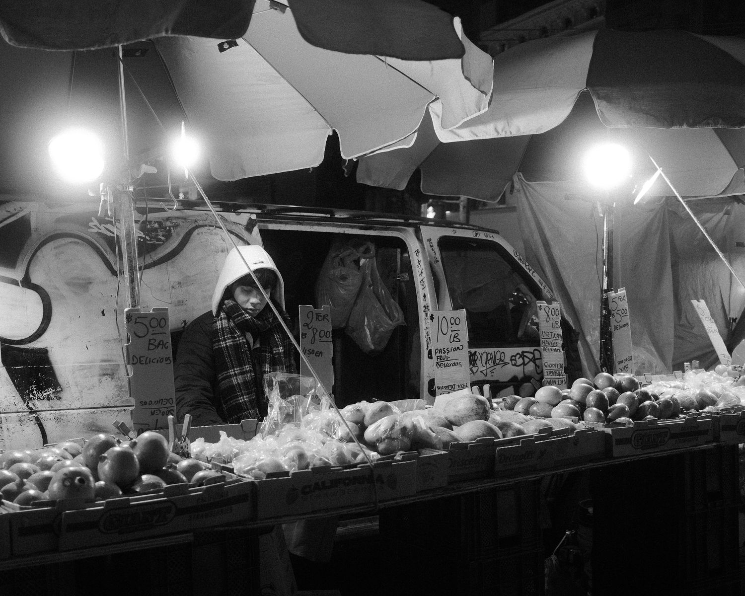 Street vendor standing behind a produce stand under bright lights and umbrellas at night.