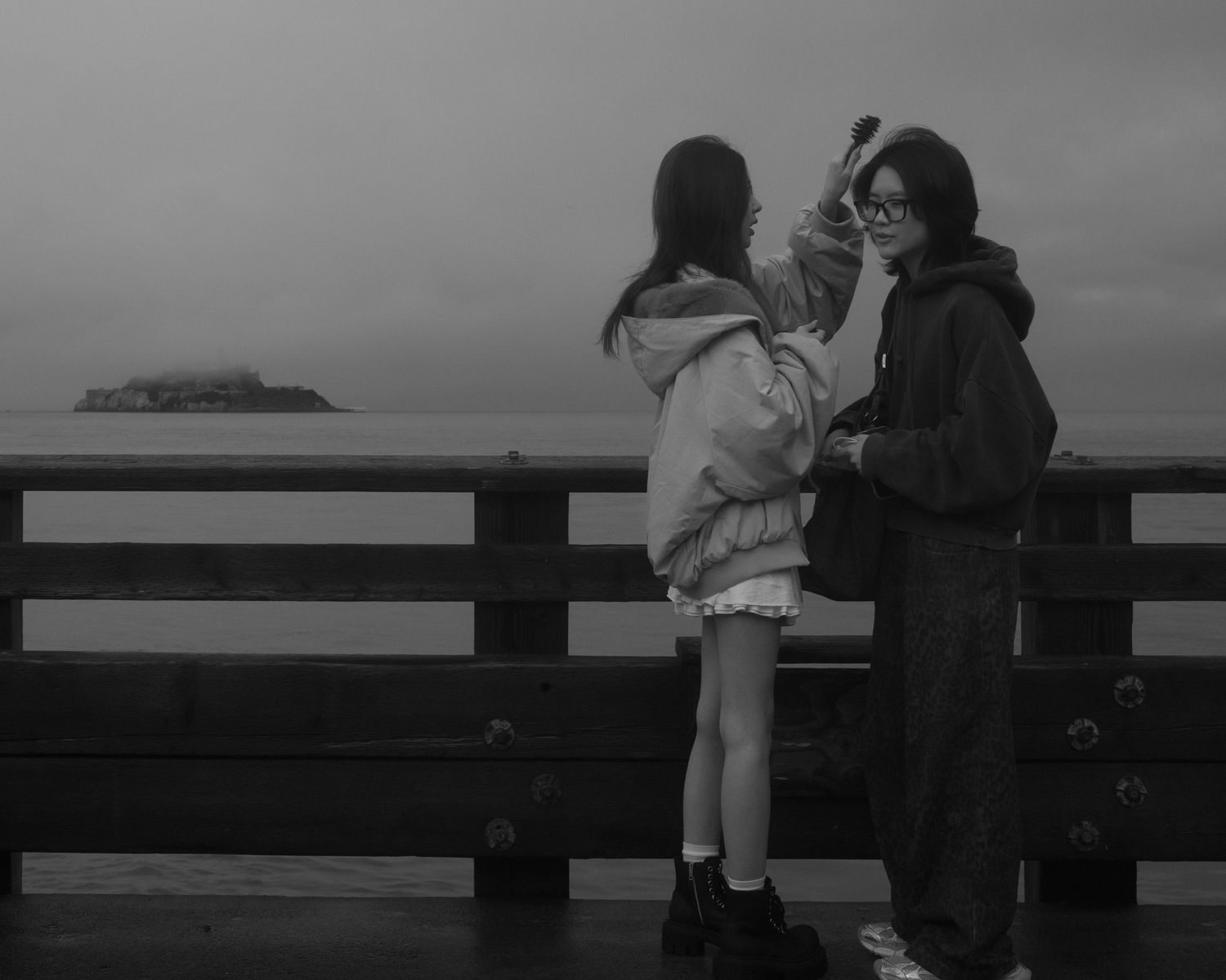 Two women standing on a pier, with one brushing the other’s hair, and an island visible in the distance.