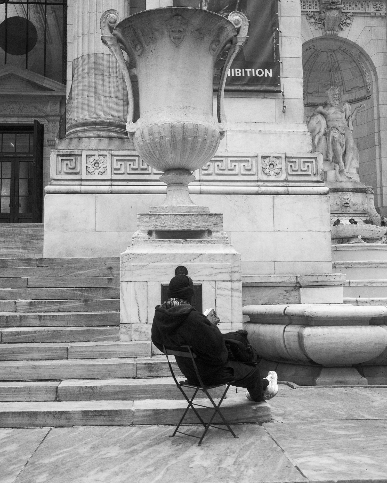 Person sitting on a folding chair reading near a large stone urn on the steps of a historic building.