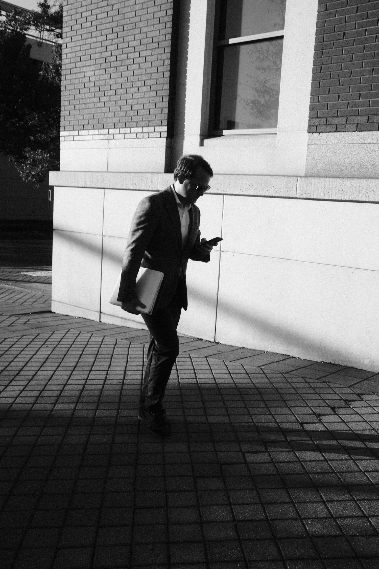 Man walking on a brick sidewalk while looking at his phone and holding a laptop.
