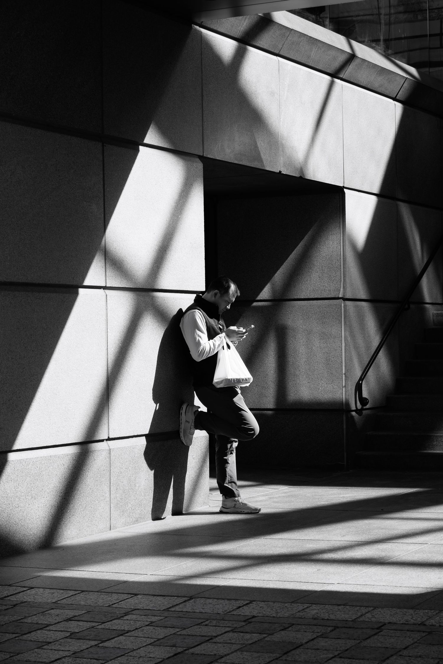 Man leaning against a large concrete wall while looking at his phone with diagonal shadows on the ground.