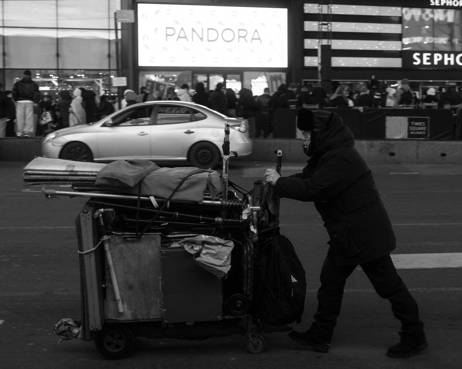 Person pushing a loaded cart across a city street with a crowd and storefronts in the background.