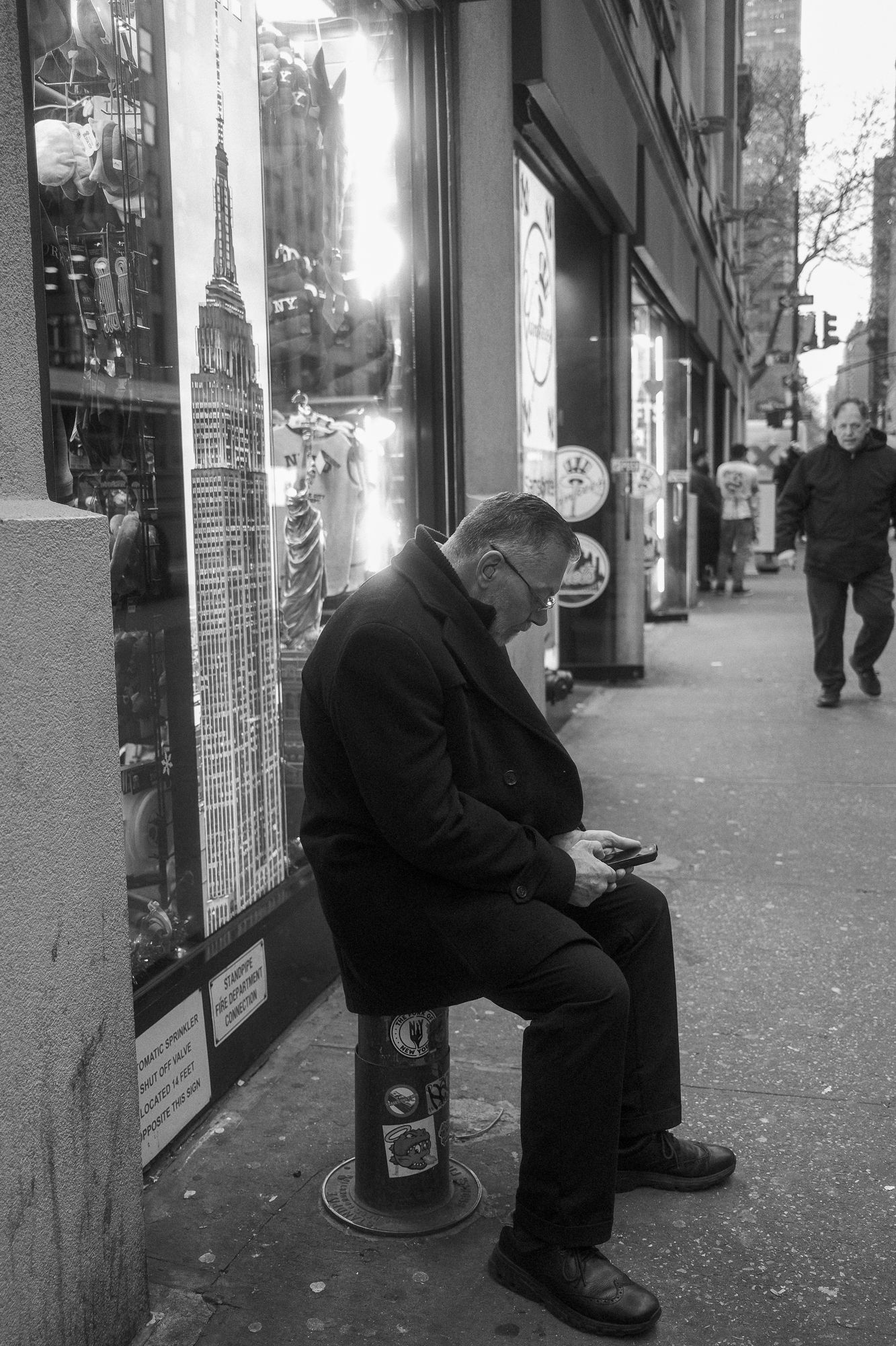 Man sitting on a small seat outside a store window while looking at his phone.