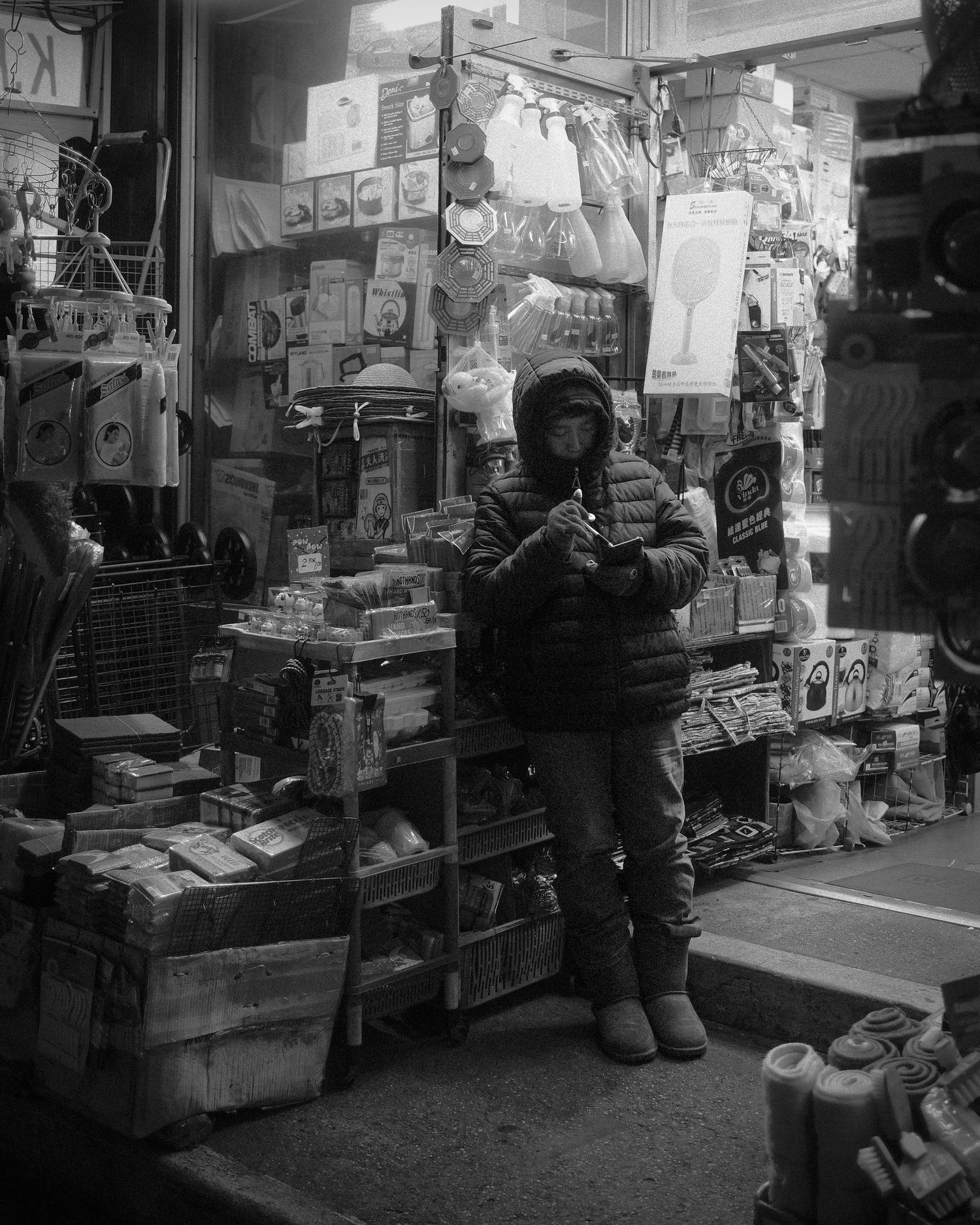 Person wearing a hooded jacket standing in front of a shop with shelves of assorted goods.