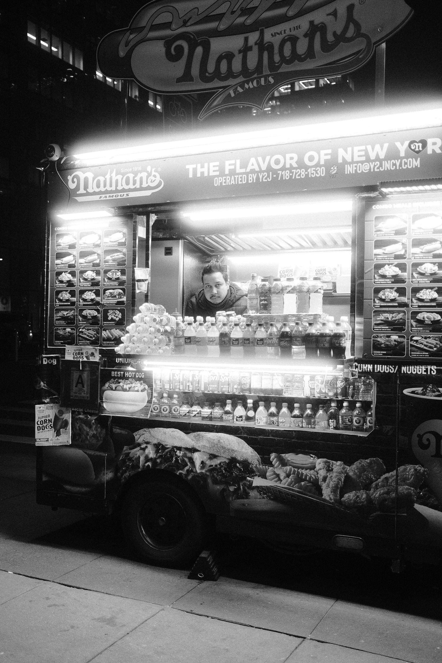 Vendor standing inside an illuminated Nathan’s hot dog cart surrounded by drinks and food items.