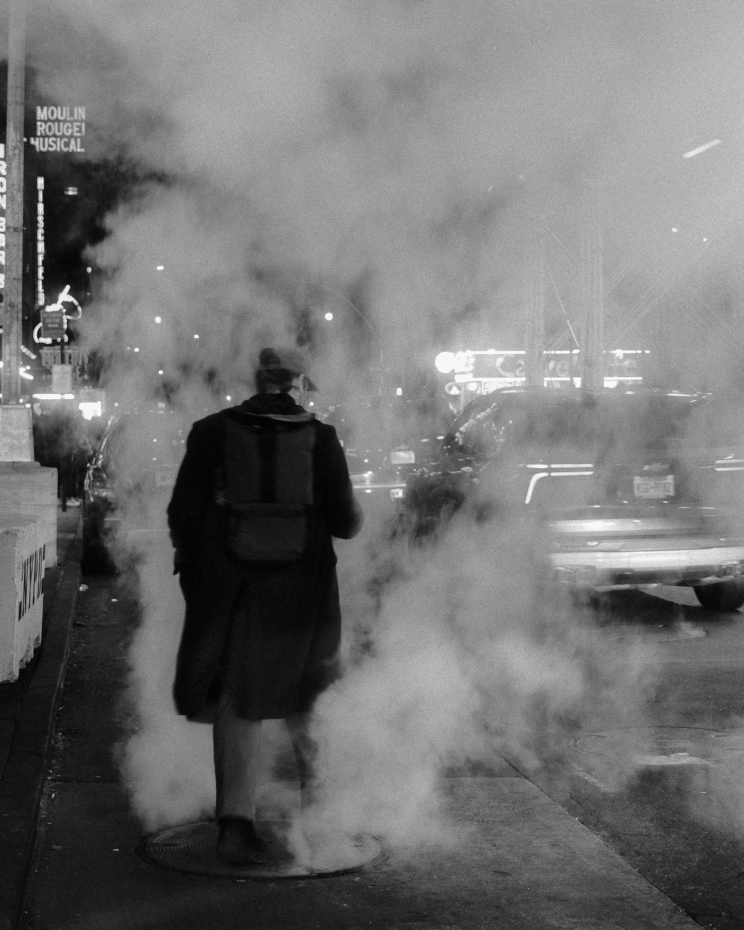 Person walking through steam coming from a street vent in a city at night.