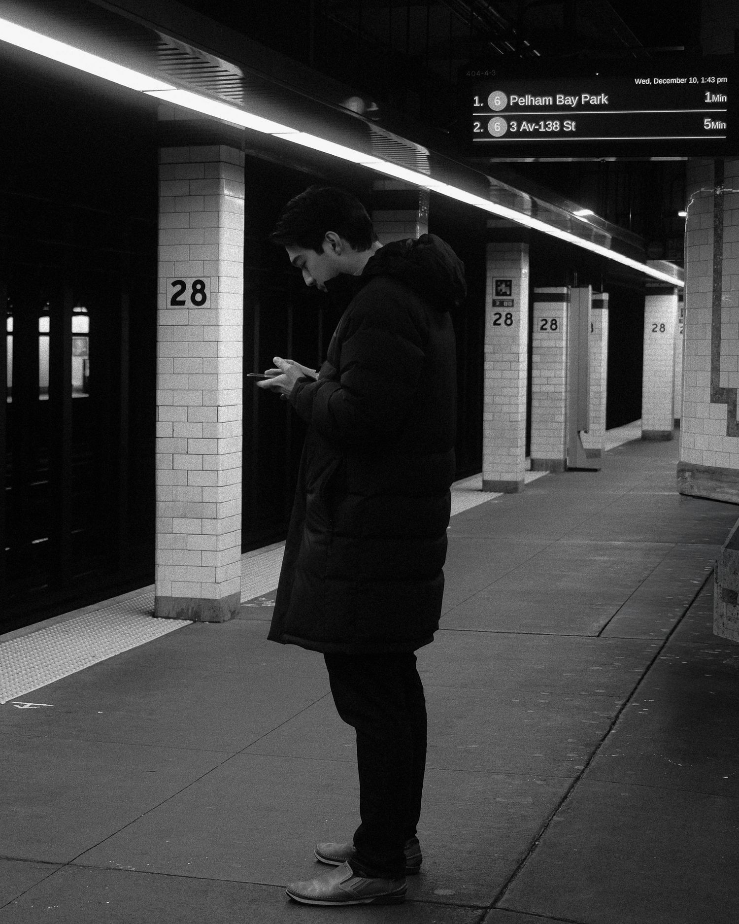 an standing on a subway platform while looking at his phone near tiled columns and overhead signs.
