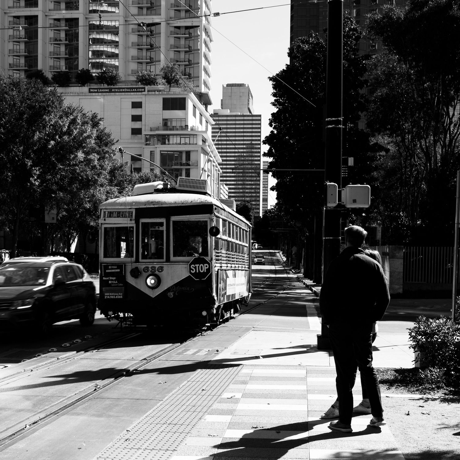 Man standing at a crosswalk watching an approaching streetcar in a downtown urban area.