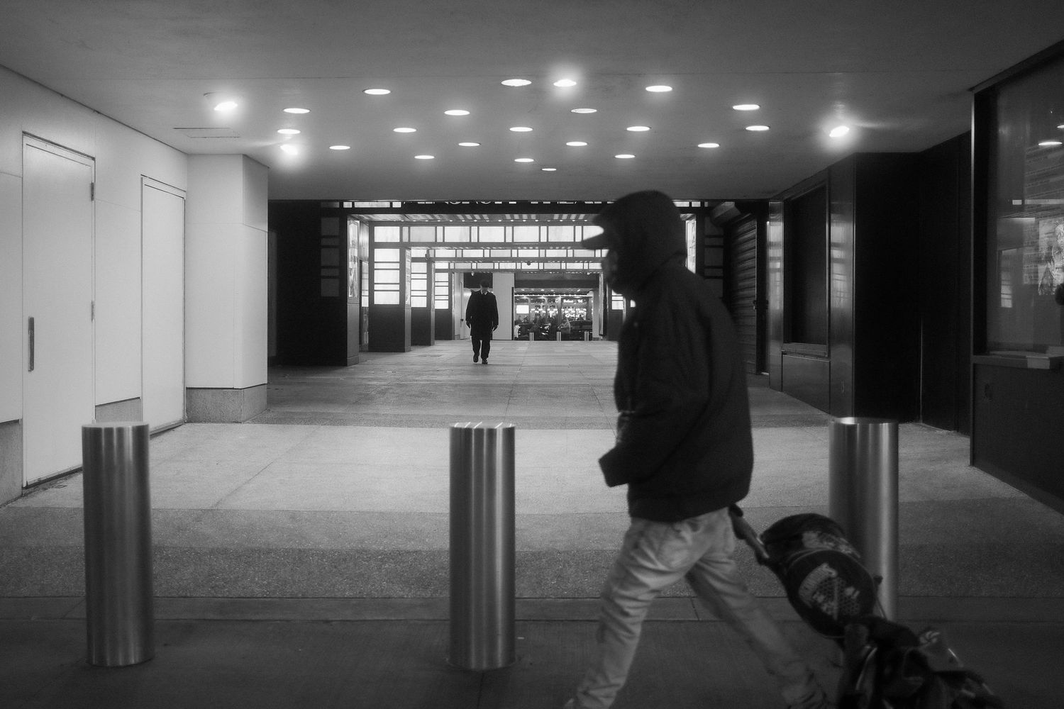 Person in a hooded jacket walking past metal bollards in a covered walkway, with another person visible in the distance.