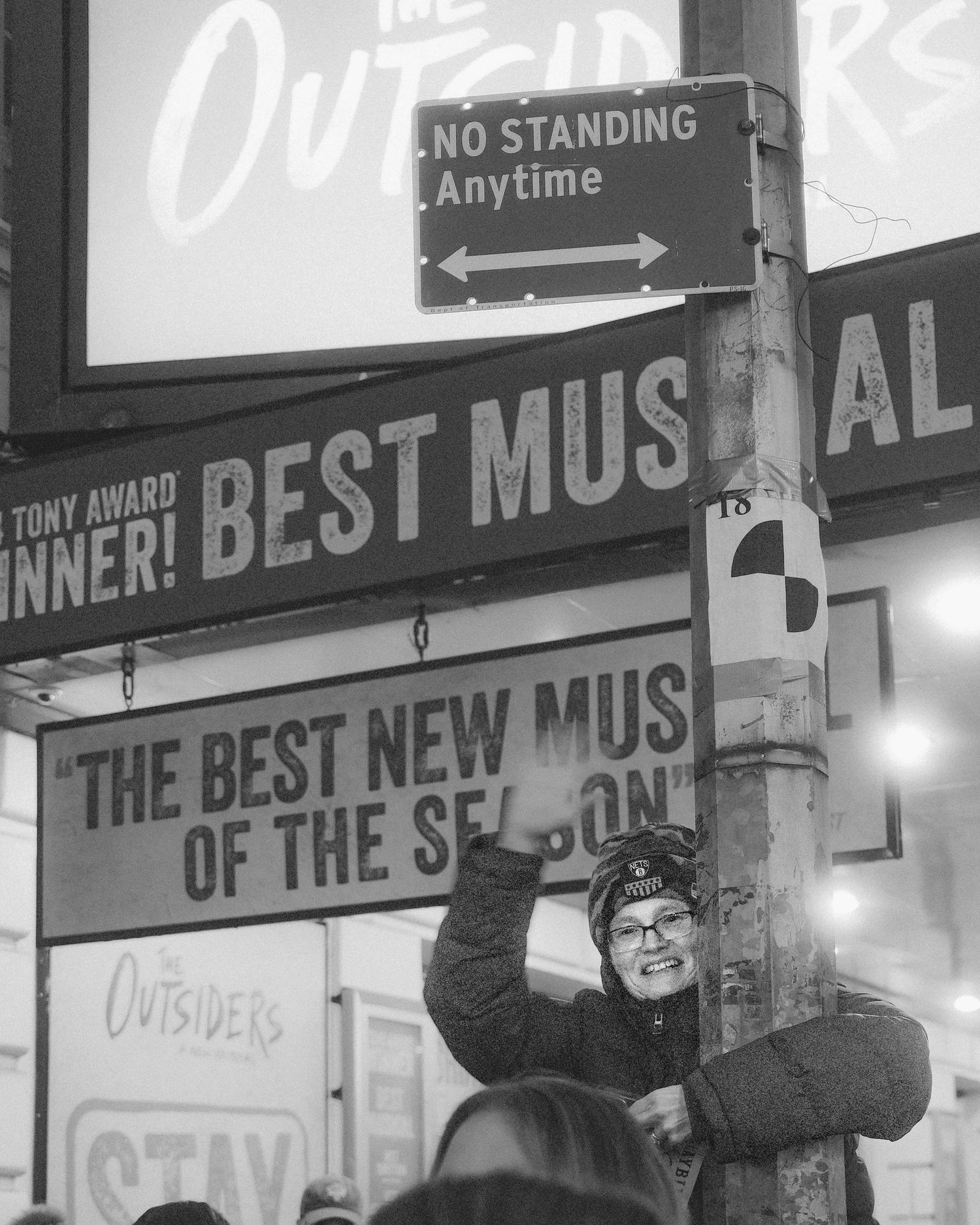 Person standing beside a pole and waving near theater signs and a “No Standing Anytime” street sign.