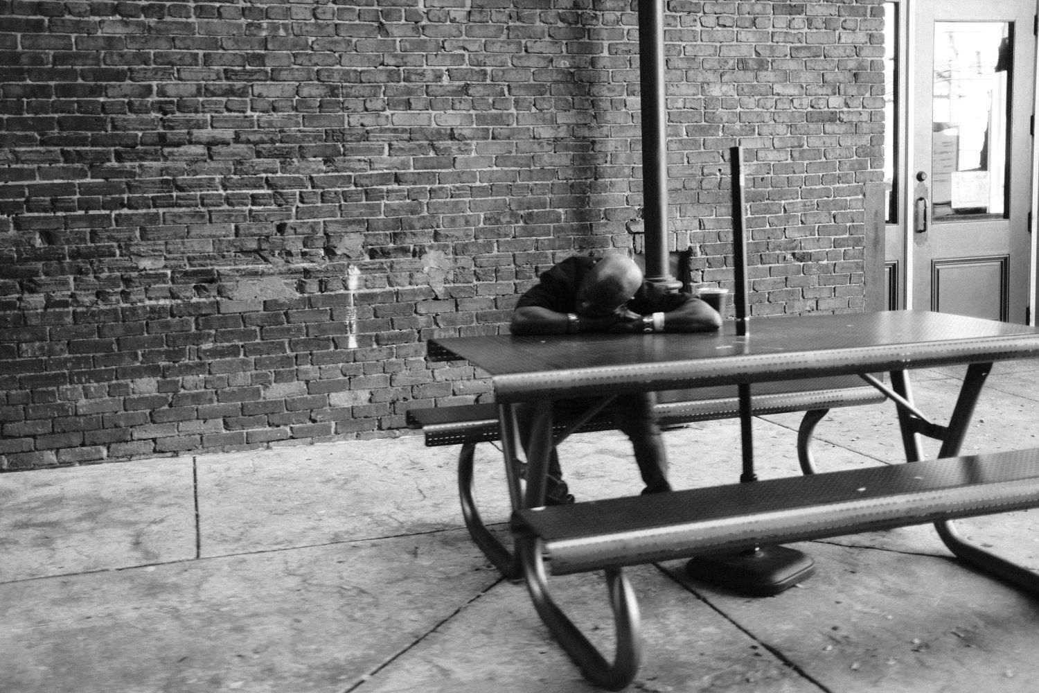 Person sitting with their head down on a picnic table next to a large brick wall.