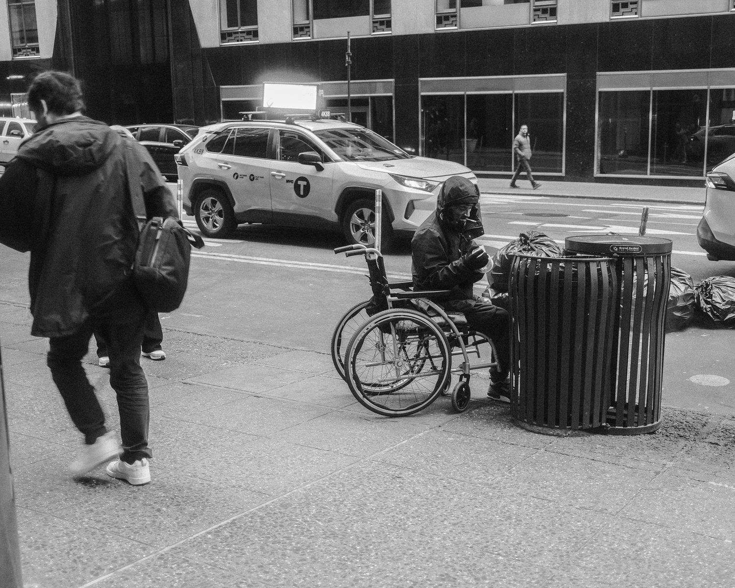 Person sitting in a wheelchair beside a trash can on a busy city sidewalk.
