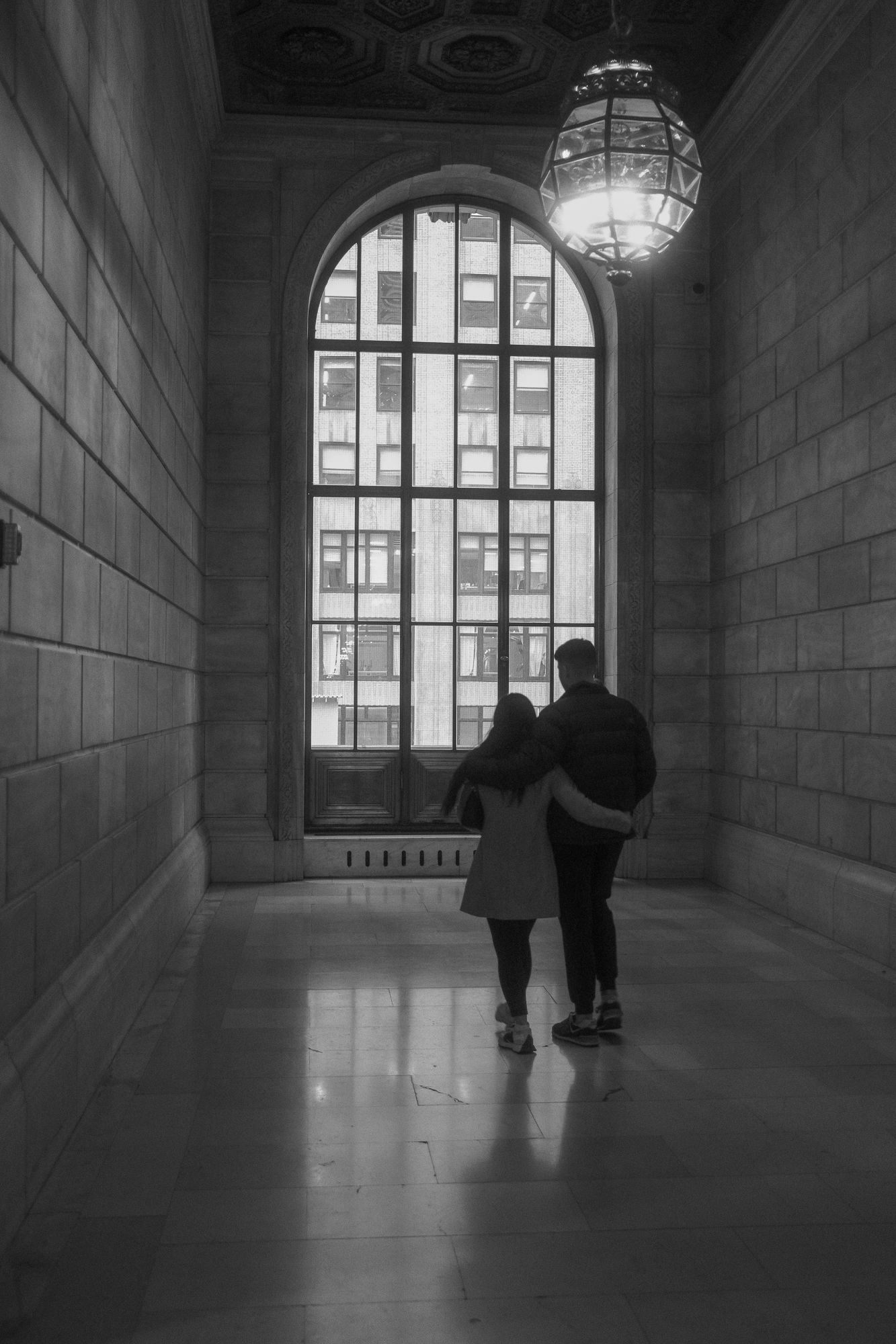 Couple walking with arms around each other in a large stone hallway toward a tall window.