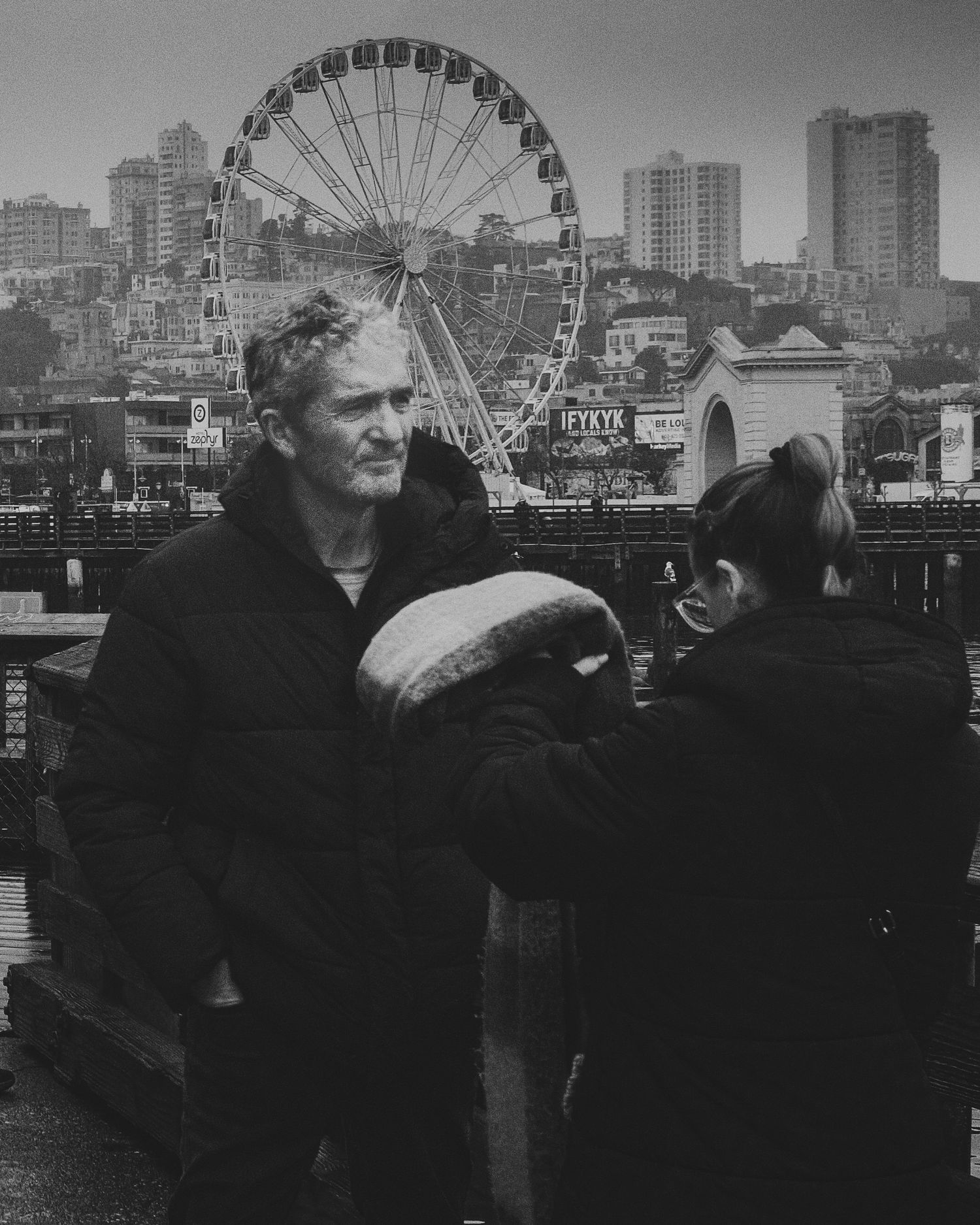 Man standing near a waterfront railing with a Ferris wheel and city skyline in the background.