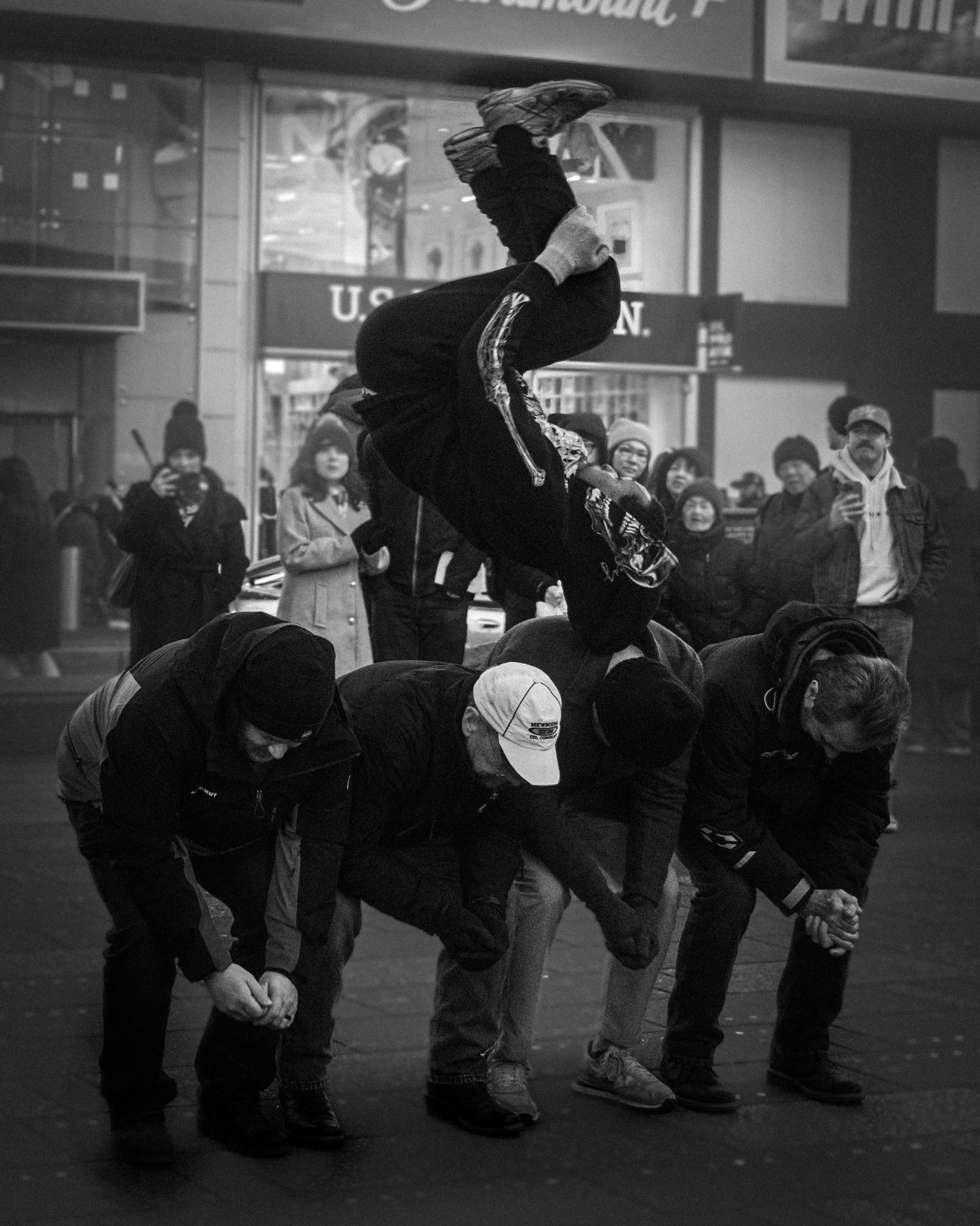 Street performer mid-air flipping over several crouched people while a crowd watches.