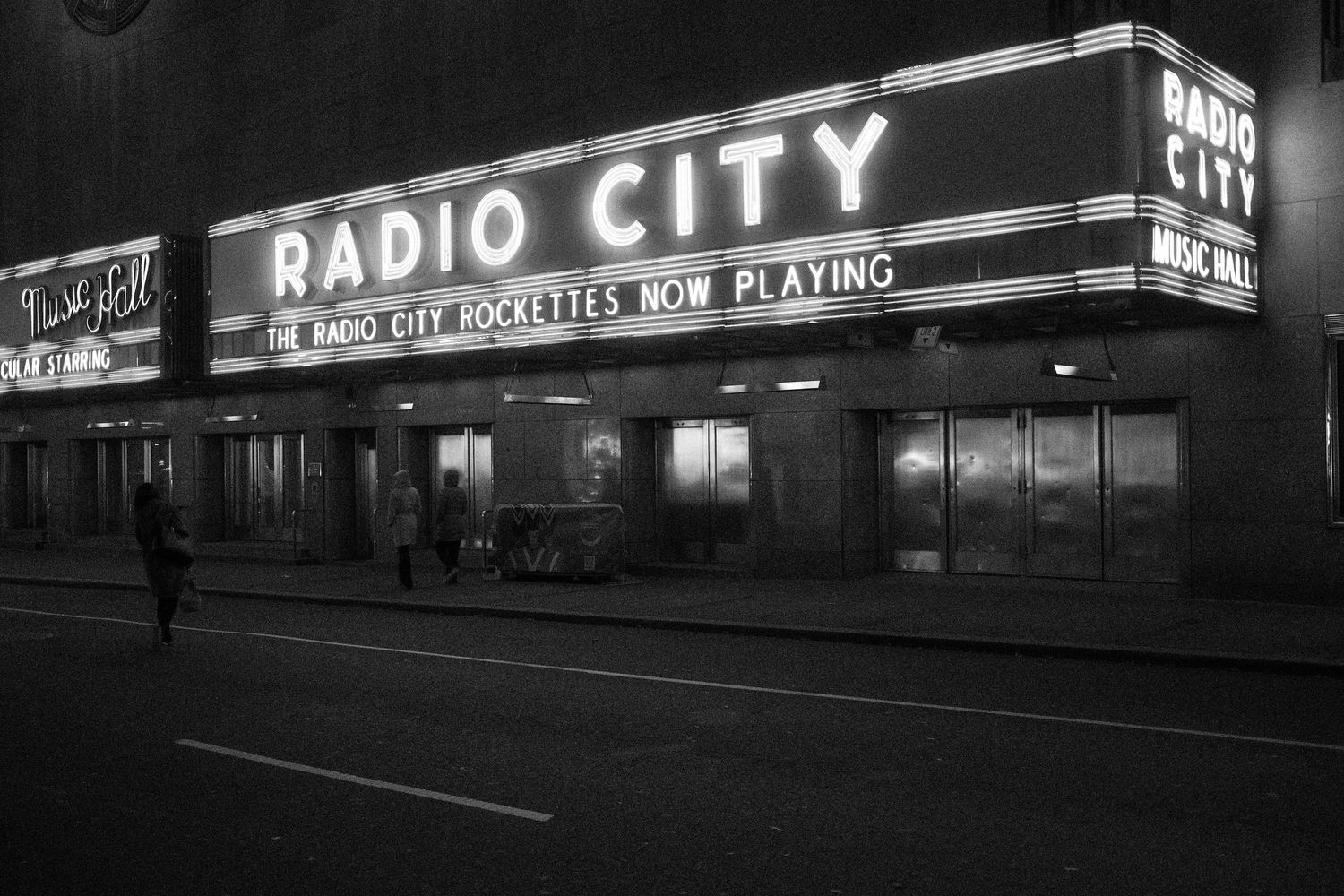 Radio City Music Hall marquee lit at night with a few pedestrians walking along the sidewalk.