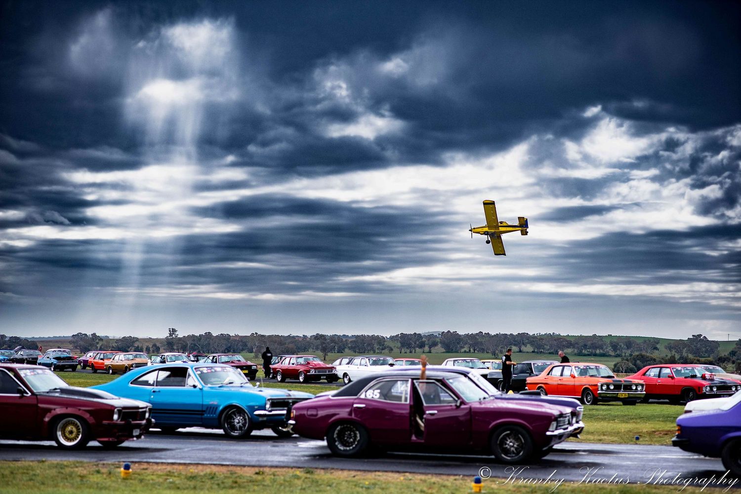 Cars line up on the Cootamundra airstrip for the Coota 400 while a plane flies overhead. Streaks of sunlight filter through storm clouds.