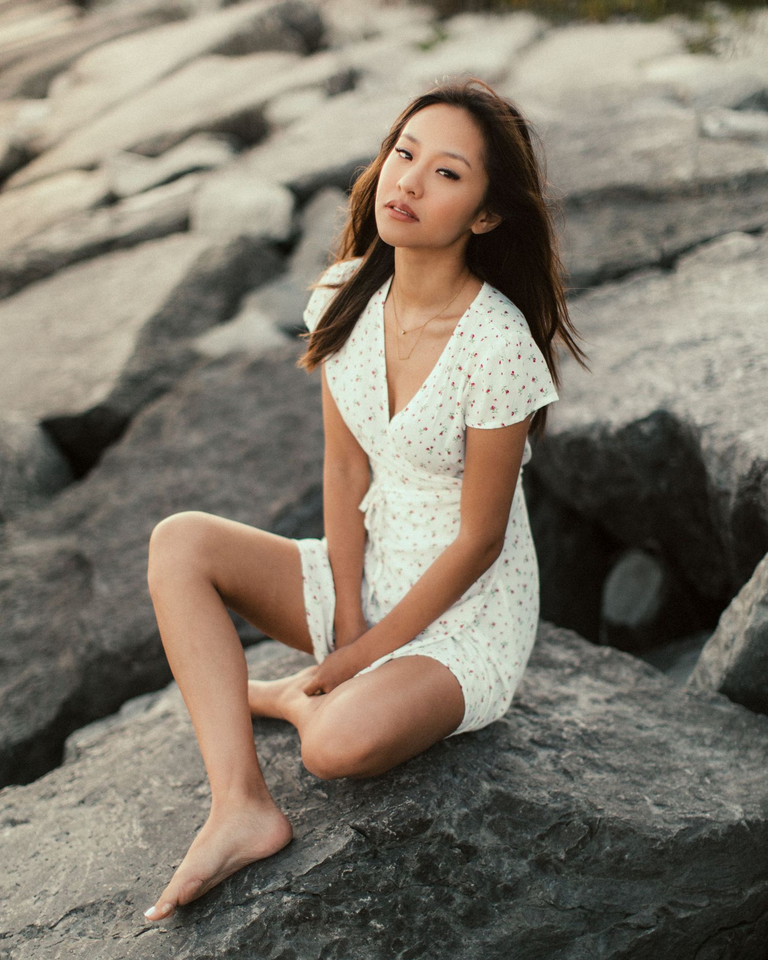 Model in white lace dress poses gracefully on rocky coastal formation at sunset.
