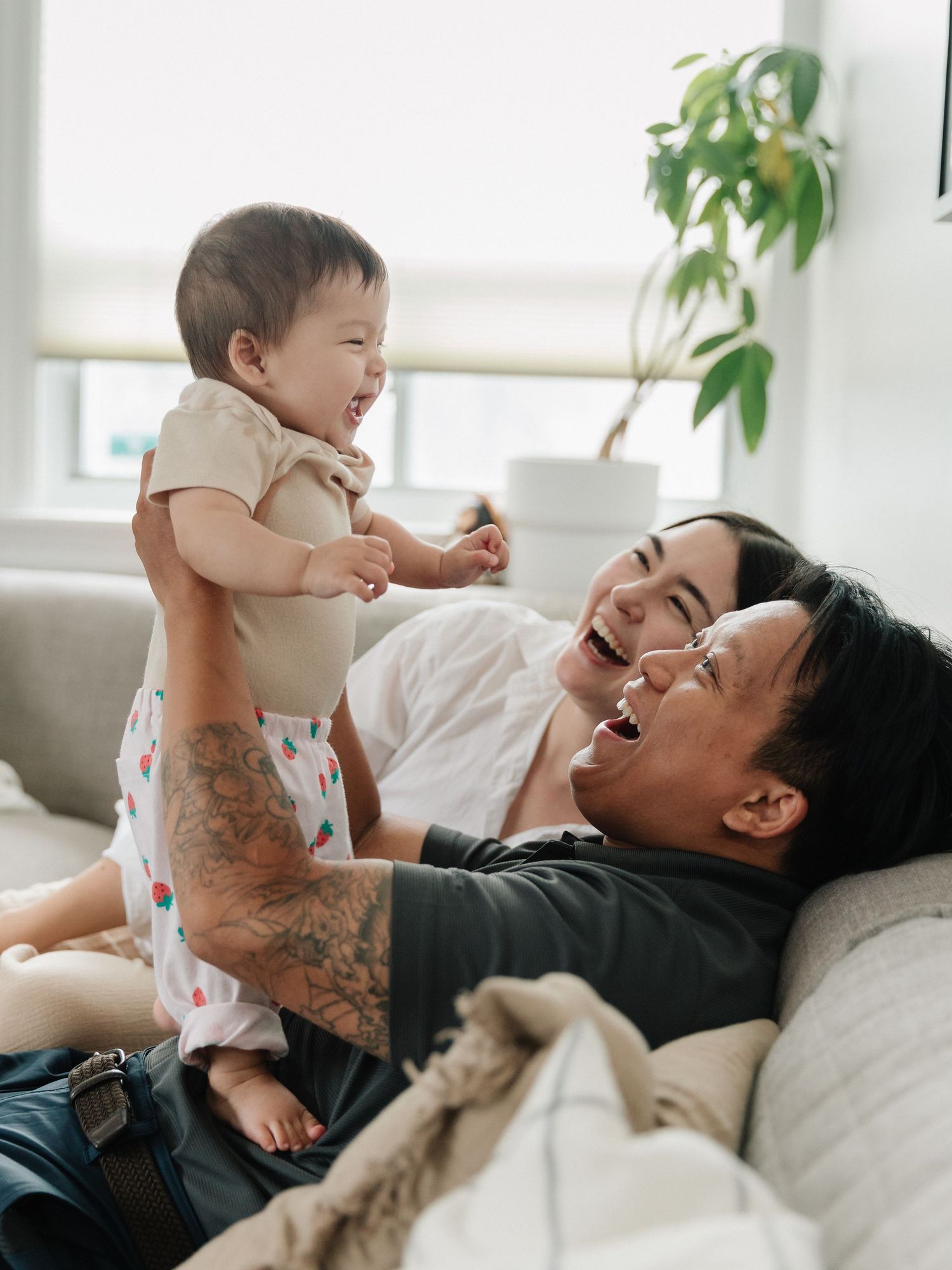 Two parents laughing joyfully with their happy baby on a cozy sofa in a bright living room.