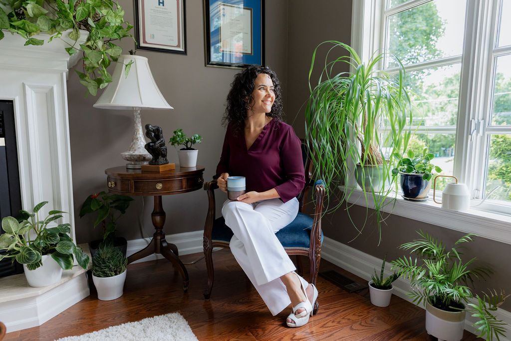 Woman relaxing in a plant-filled living room, sitting in a chair by a window, holding a coffee mug.