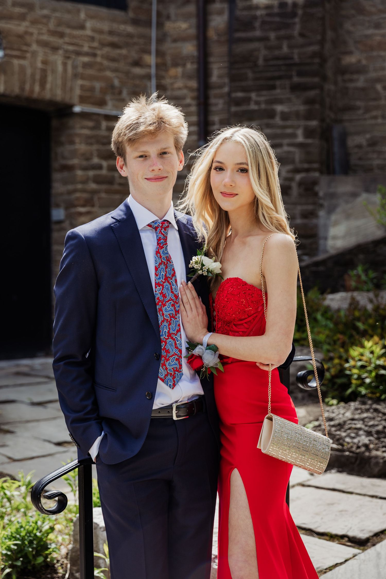 A young couple dressed for prom, man in navy suit with red tie, woman in red strapless gown, posing outdoors.