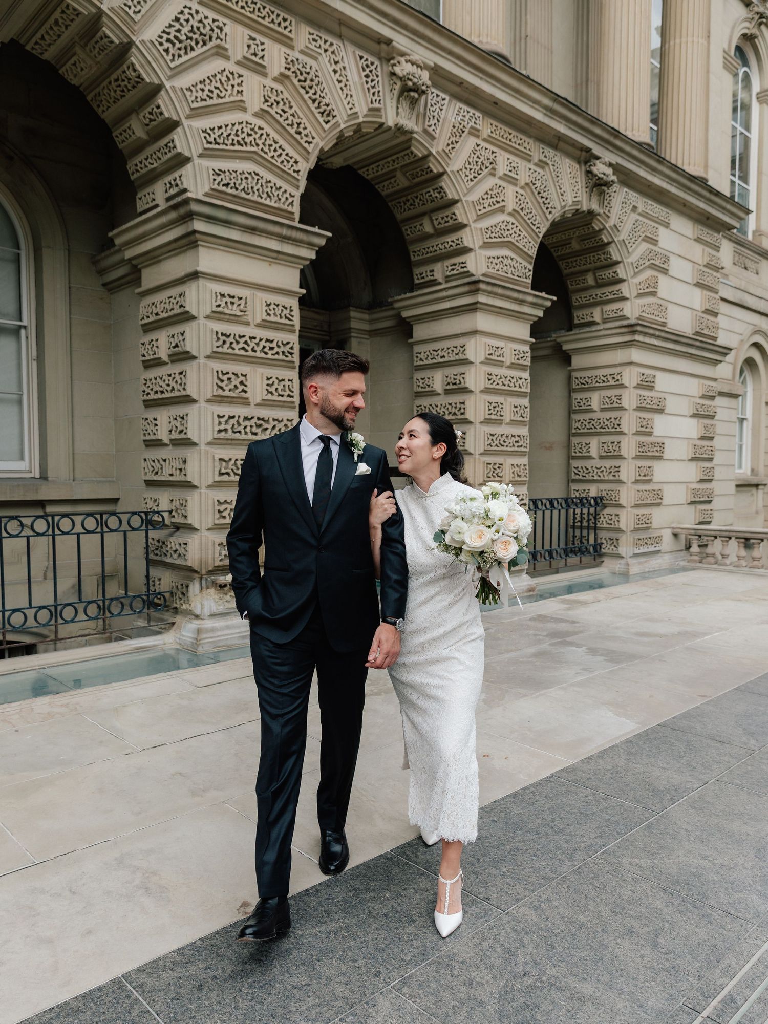 A couple in wedding attire walks outside a grand stone building, the bride holding a white bouquet in a midi dress.