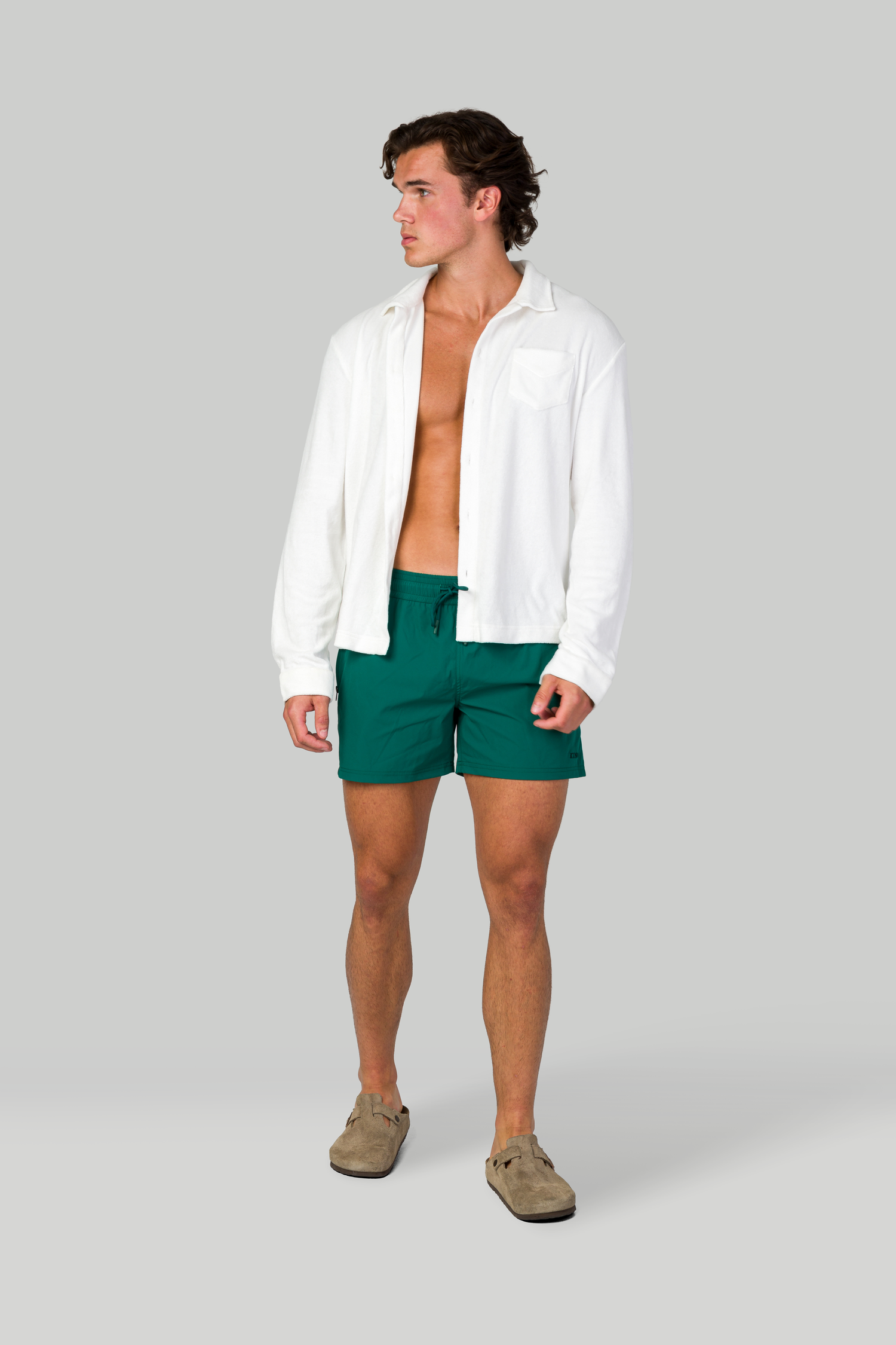 Open white shirt with green swim shorts and beige loafers against gray backdrop.