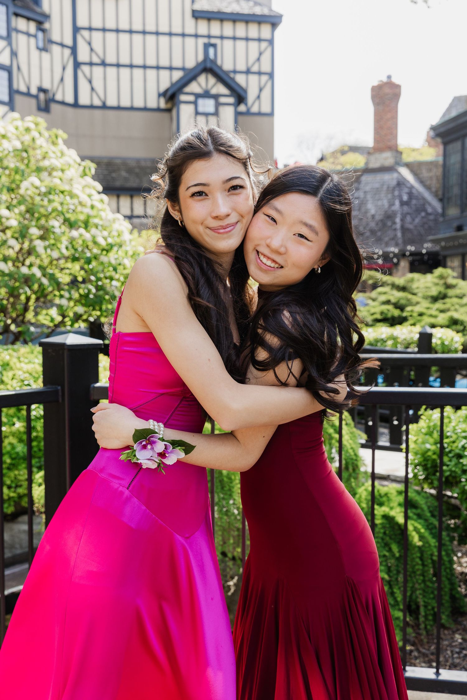 Two young women in pink and red prom dresses hugging outdoors near a garden railing.