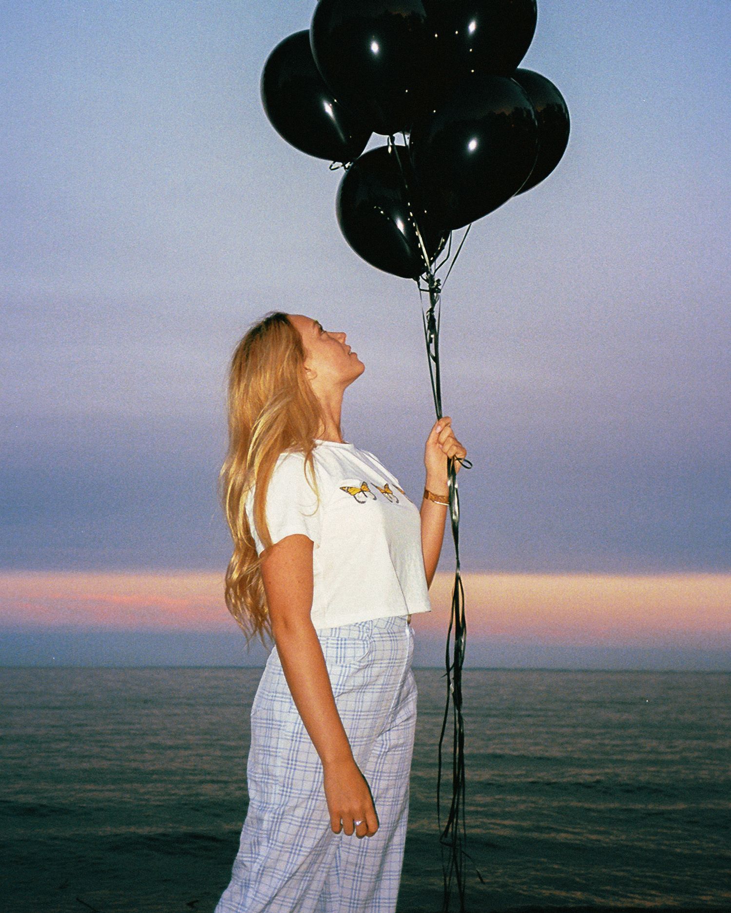 Person in white dress holding black balloons against sunset ocean backdrop.