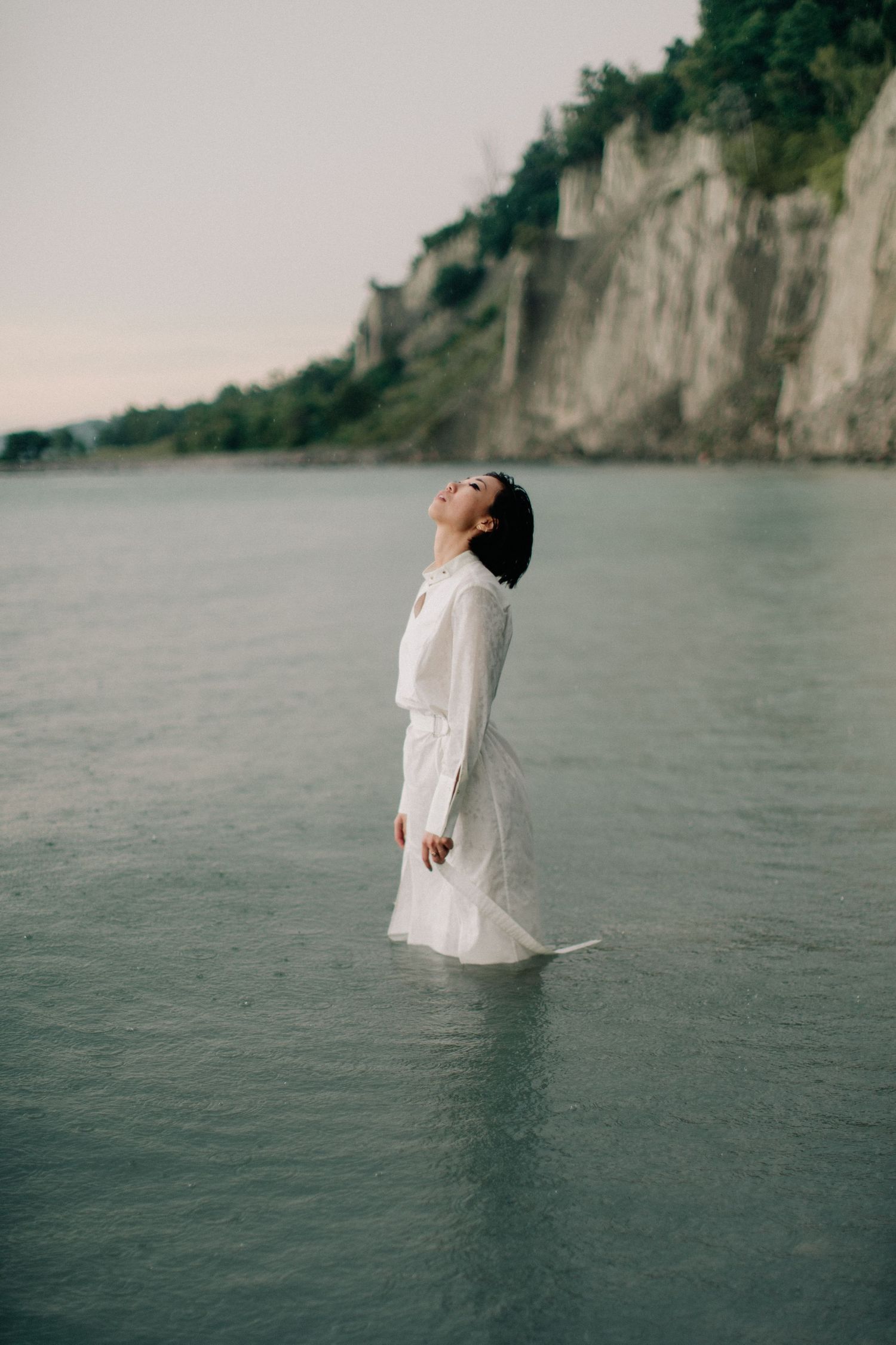Woman in white dress standing in calm water near a rocky cliff, head tilted back in serene pose.