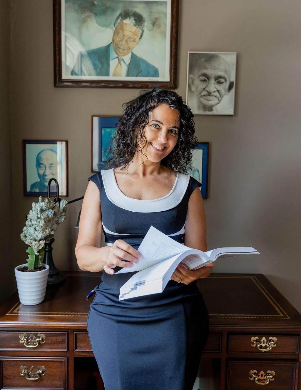 Professional woman in navy dress holding documents, standing by wooden desk with portrait paintings on wall behind her.