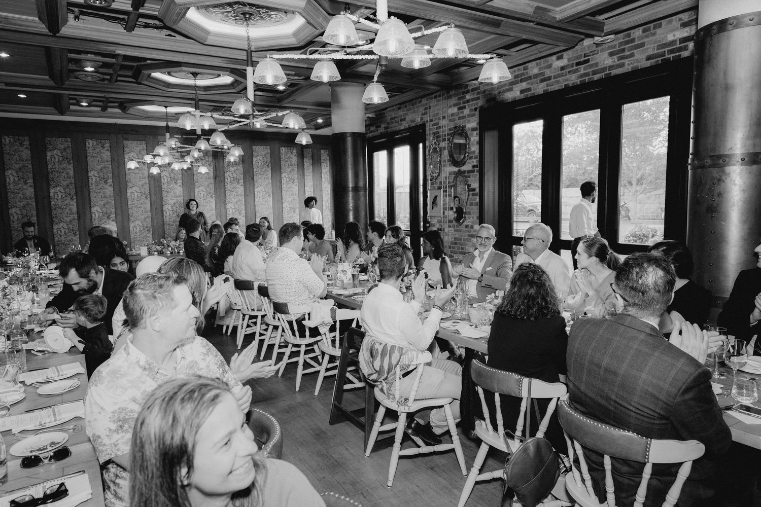 Black and white photo of guests dining at elegantly set tables in a rustic brick-walled restaurant with pendant lights.