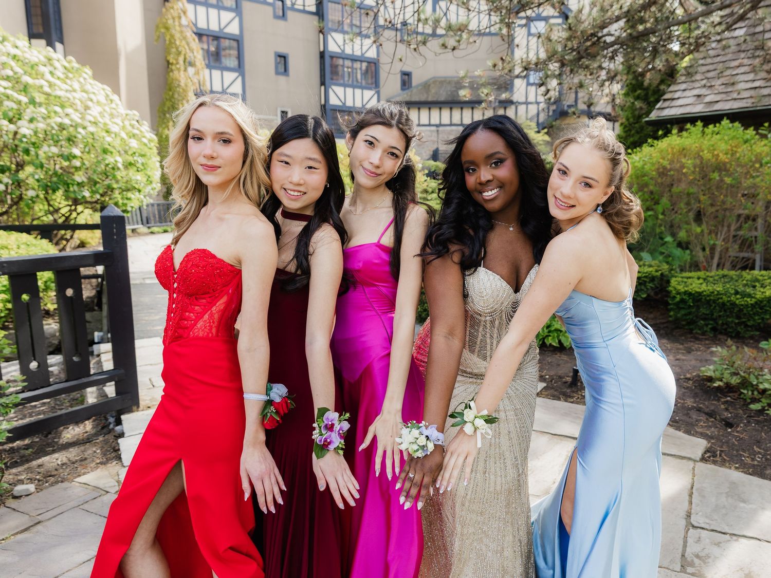 Five young women in elegant prom dresses pose outdoors, wearing floral wrist corsages in vibrant colors.