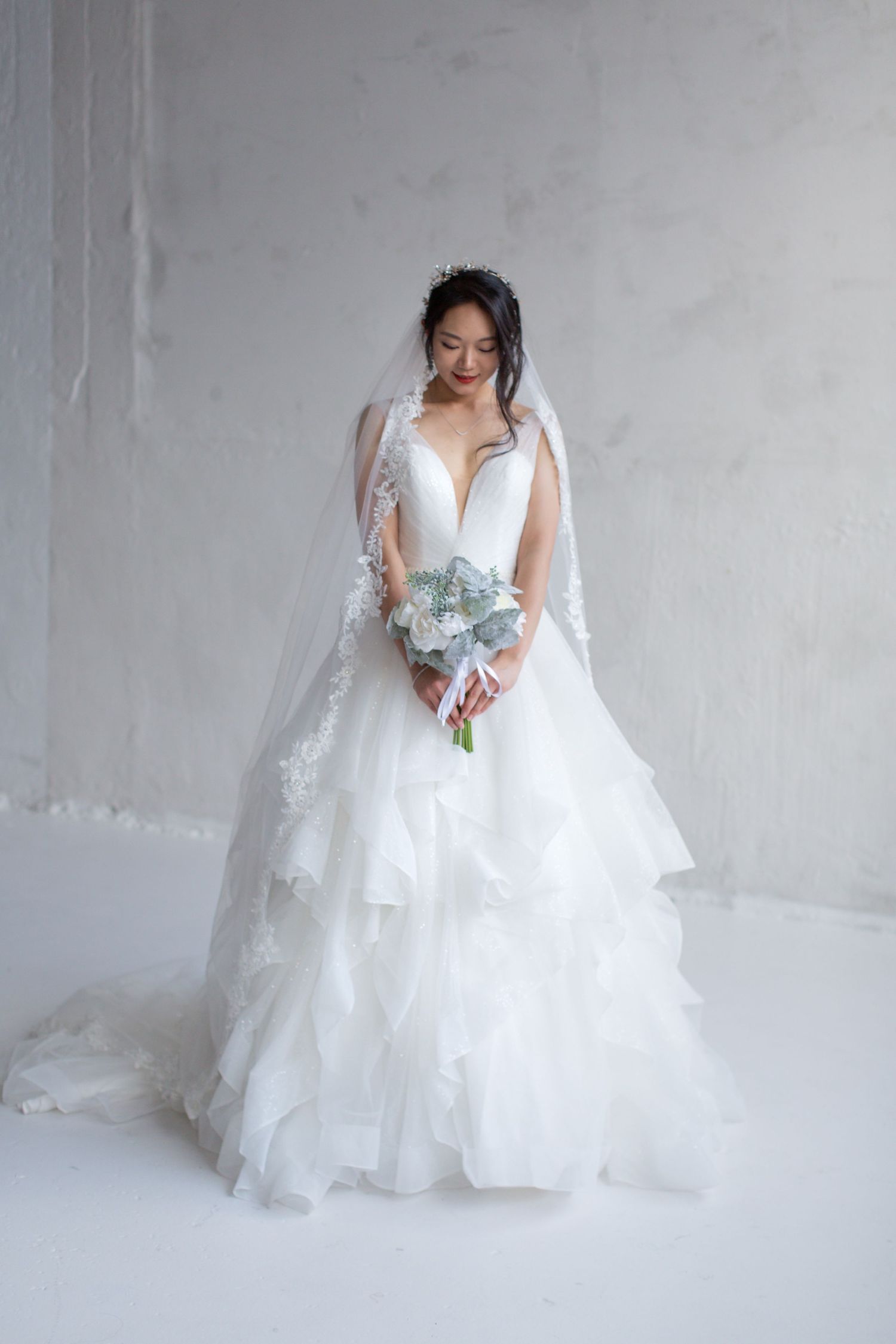 Bride in an elegant white layered wedding gown with veil holds a blue bouquet against a light backdrop.