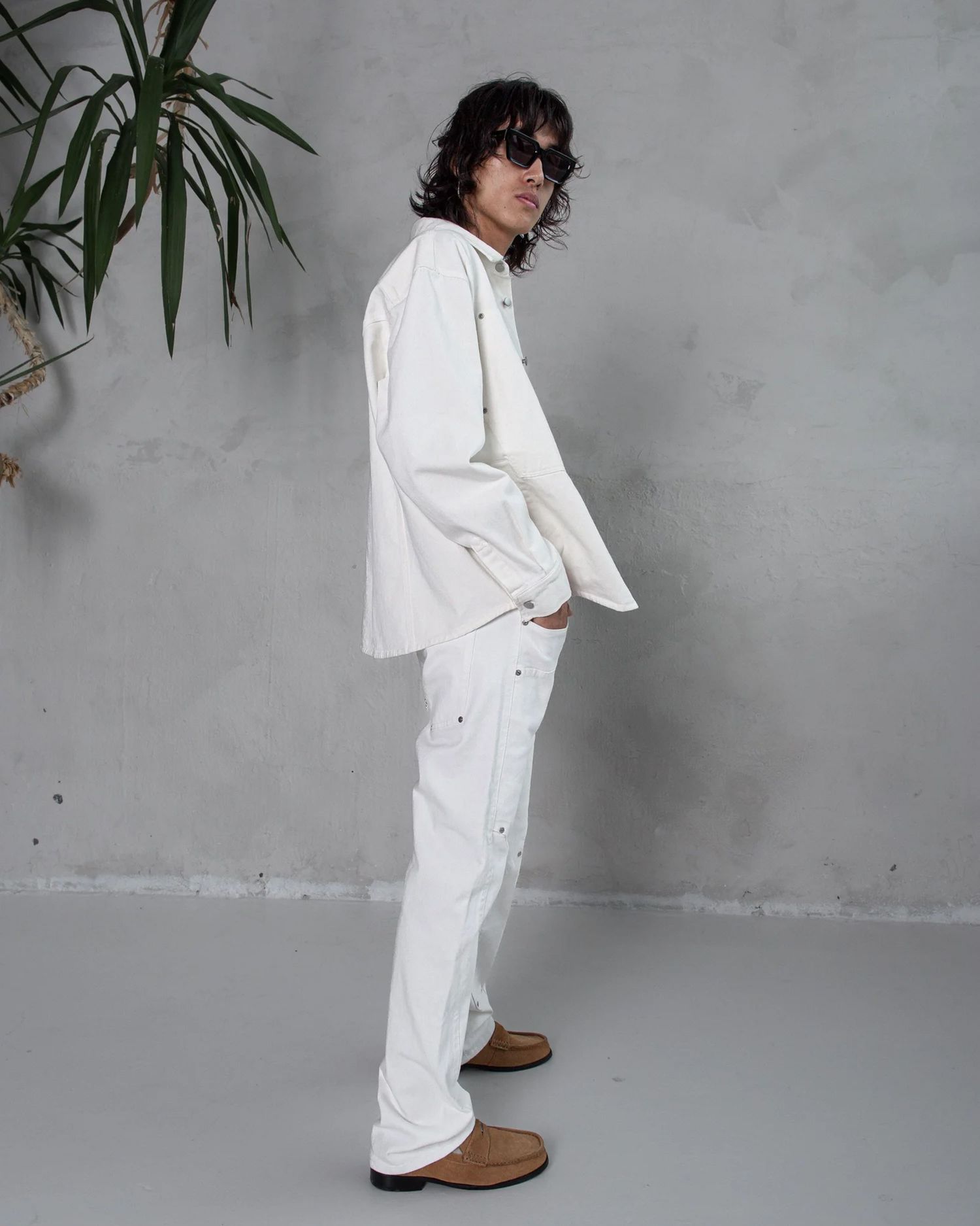 Fashion shoot featuring all-white outfit with brown boots against grey wall with plant.