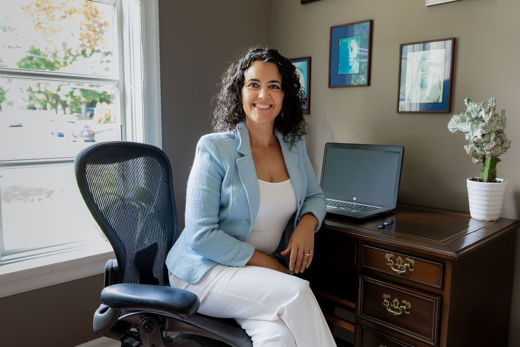 Woman in light blue blazer and white pants sitting confidently at a wooden desk with laptop in a modern office.