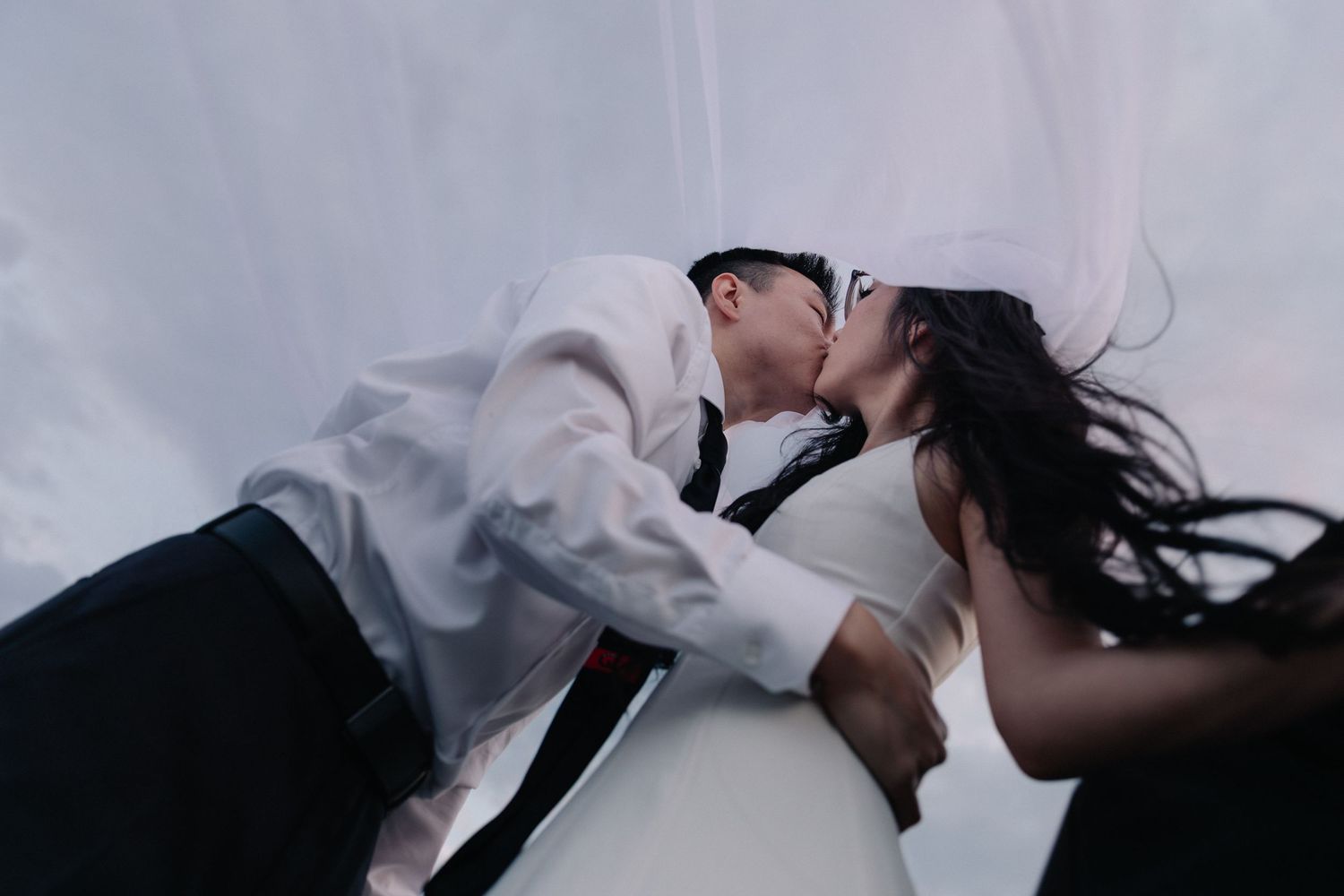 A couple shares a romantic kiss during their wedding, with the bride's veil flowing in the wind against a white backdrop.
