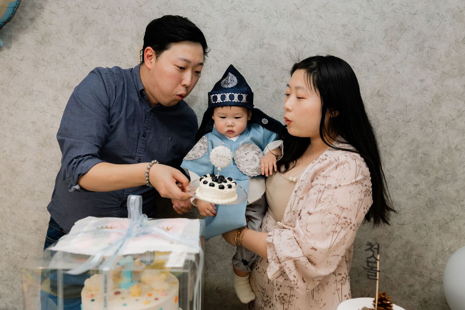 Asian family celebrating baby's first birthday with a small cake, parents blowing candles together.