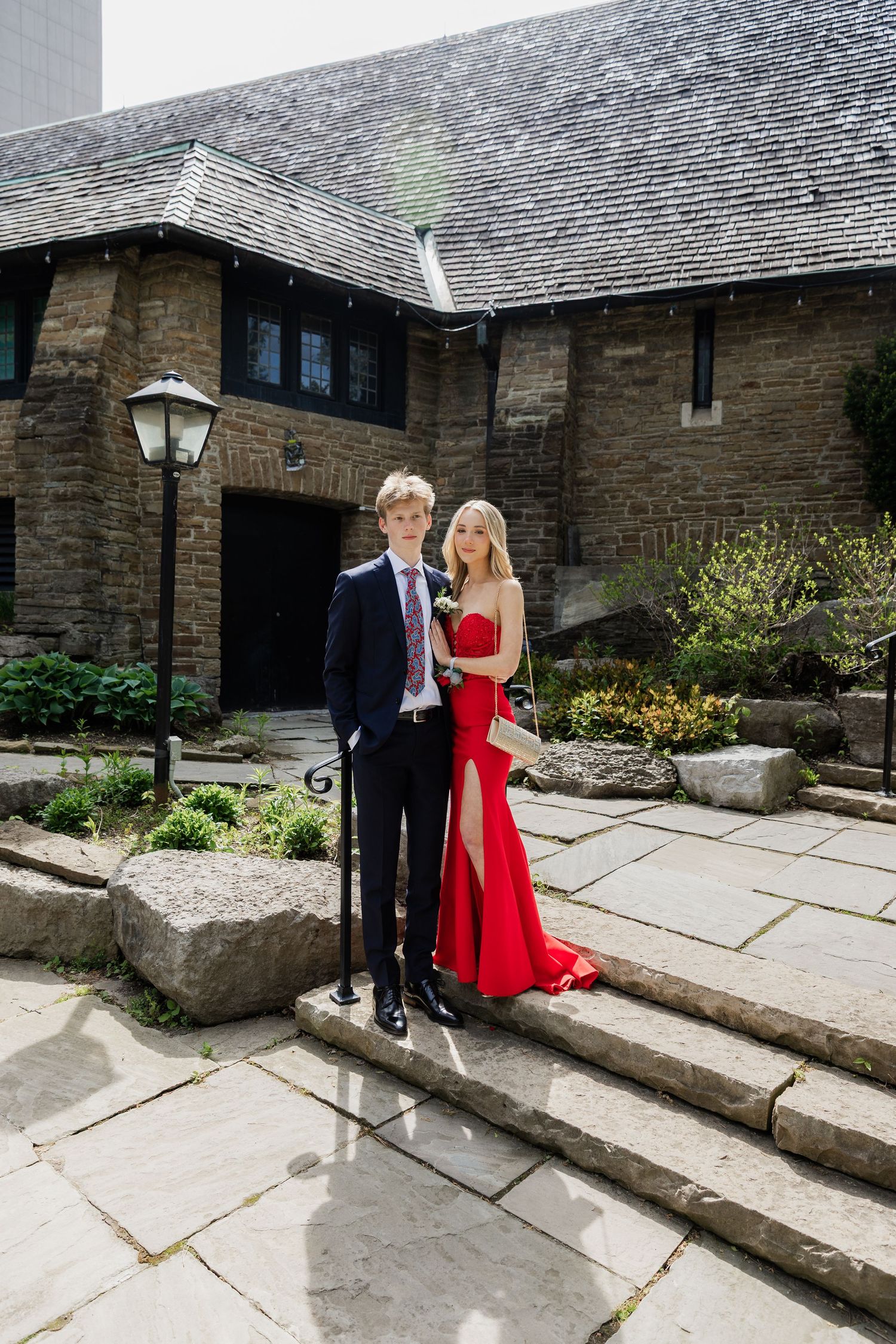 A young couple dressed in prom attire, a red gown and navy suit, posing on stone steps outside a historic building.