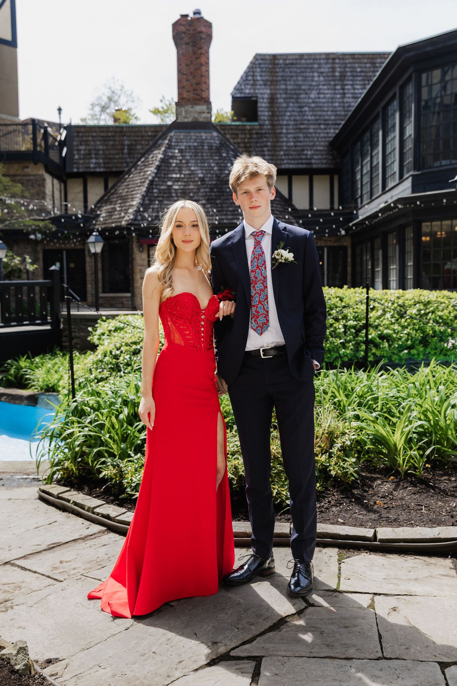 A young couple dressed in prom attire, woman in red gown and man in navy suit, posing in a garden courtyard.
