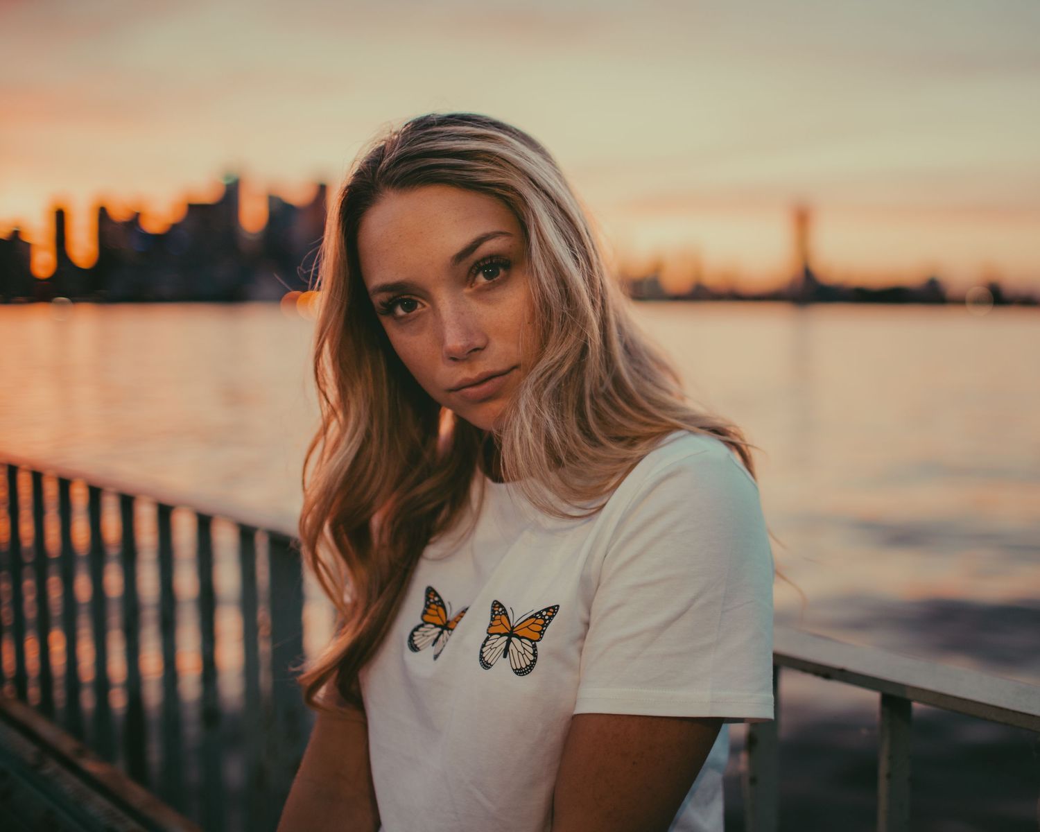 Sunset portrait by water with butterfly graphic t-shirt and city skyline backdrop.